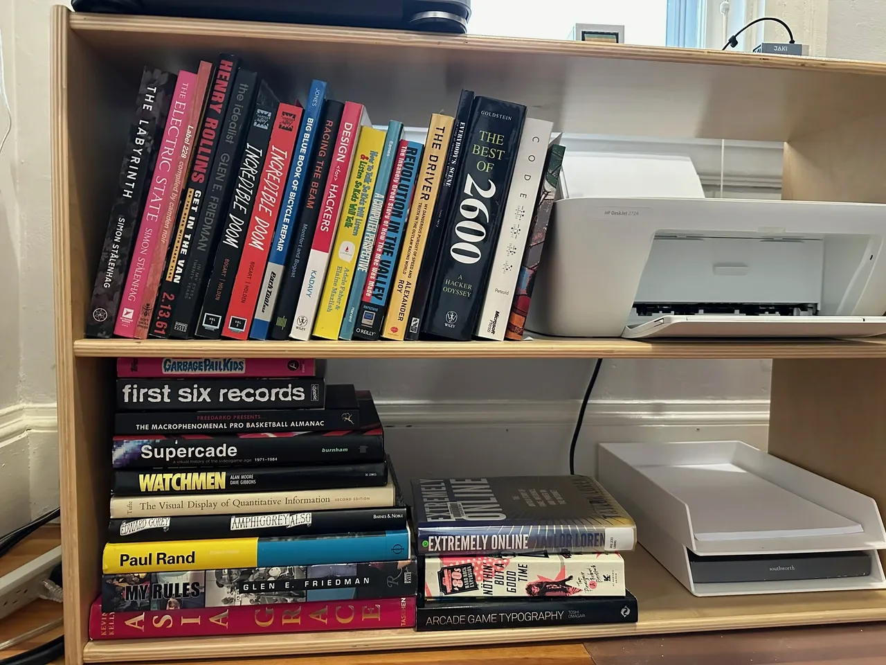 A bookshelf with a varied collection of books ranging from design to basketball almanacs and a white printer on the second shelf, situated next to a window