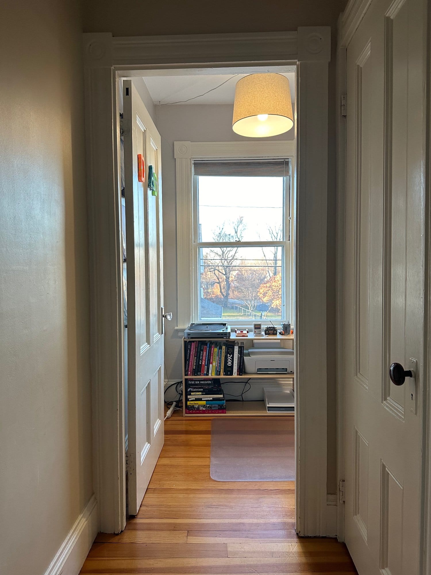 View from a hallway into a sunlit home office with a bookshelf, desk and chair by the window, and a hanging light fixture above