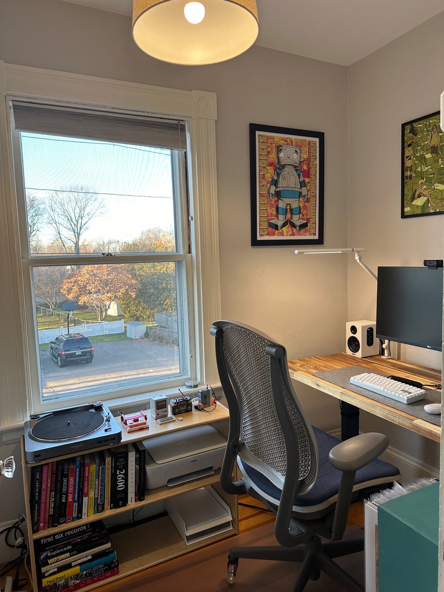 A home office with a record player and bookshelf under the window, ergonomic chair, and desk with computer and speakers, with a view of the outdoors and framed artwork on the walls