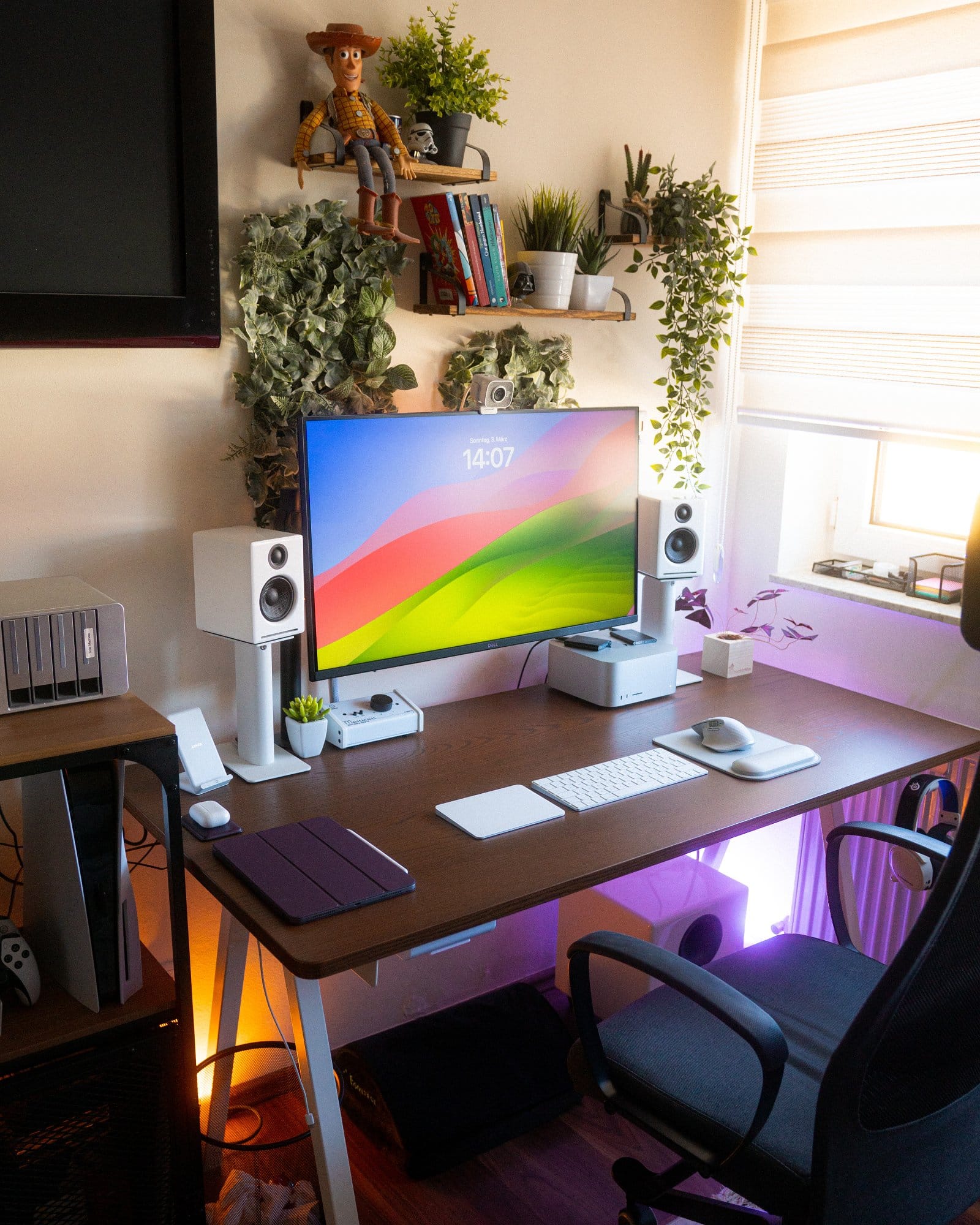 An inviting home office space with a vibrant monitor on a desk, surrounded by tech gadgets, a game console, speakers, and greenery, all under a warm light