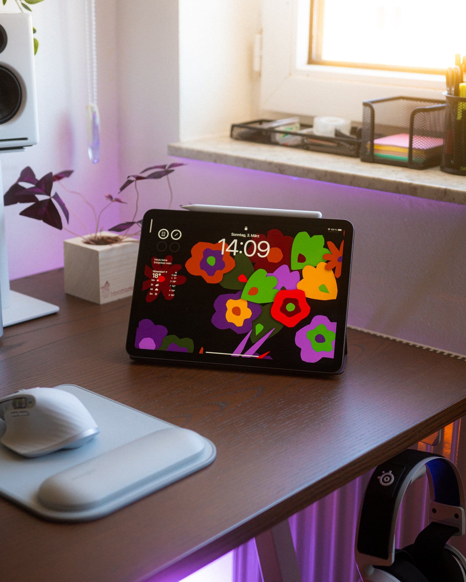 A tablet with a colourful floral display stands on a desk near an ergonomic mouse and mouse pad, with a backdrop of houseplants and office supplies