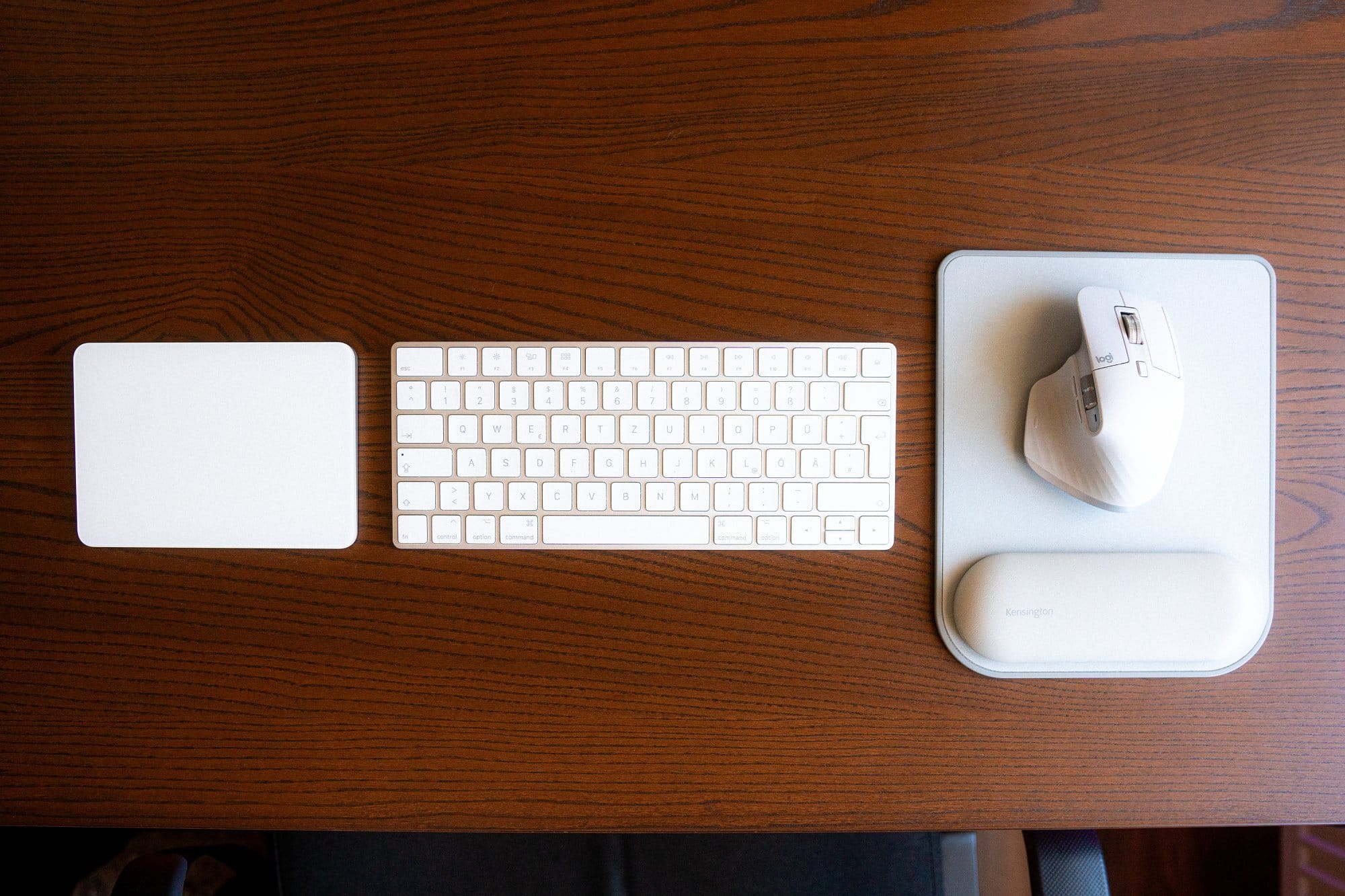 A top-down view of a workspace with a white keyboard, a trackpad, an ergonomic mouse on a mouse pad, all neatly arranged on a wooden desk