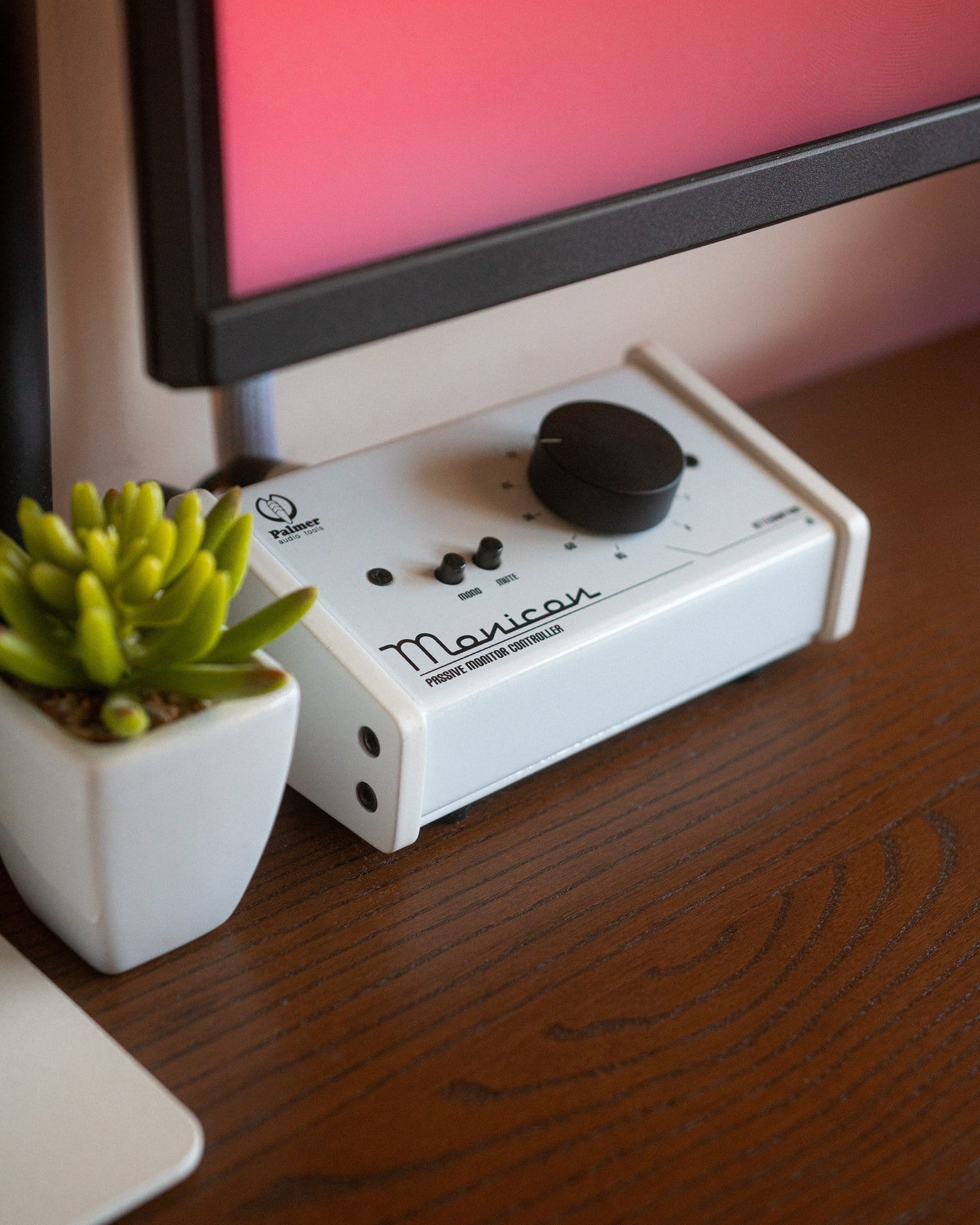 A close-up view of a desk corner showing a succulent plant in a white pot, part of a trackpad, and a monitor controller with knobs and buttons, adjacent to a computer monitor with a pinkish screen