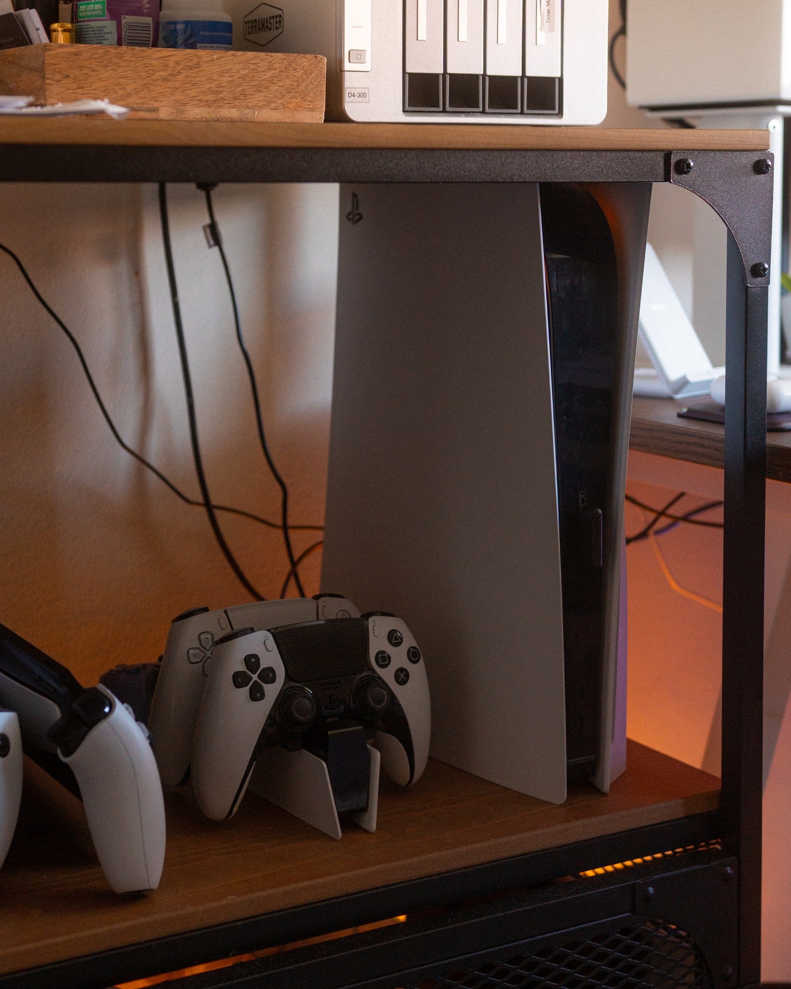 A gaming console PlayStation 5 stands upright next to its controllers on a shelf beneath a desk, with wires visible in the background and a soft glow of light
