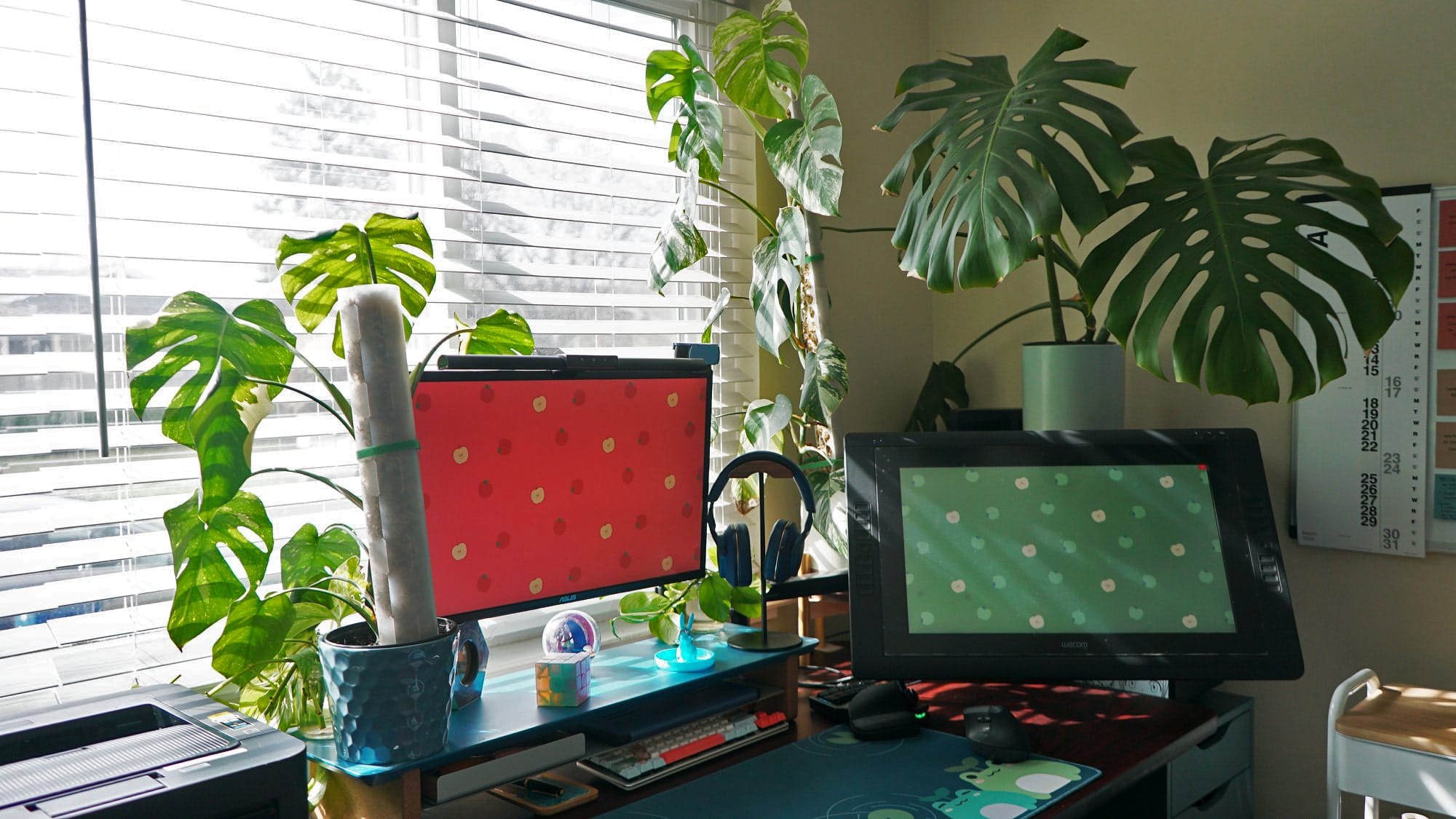 A well-lit home office featuring a large monitor, a graphic tablet, an ergonomic chair, and a variety of plants and stationery