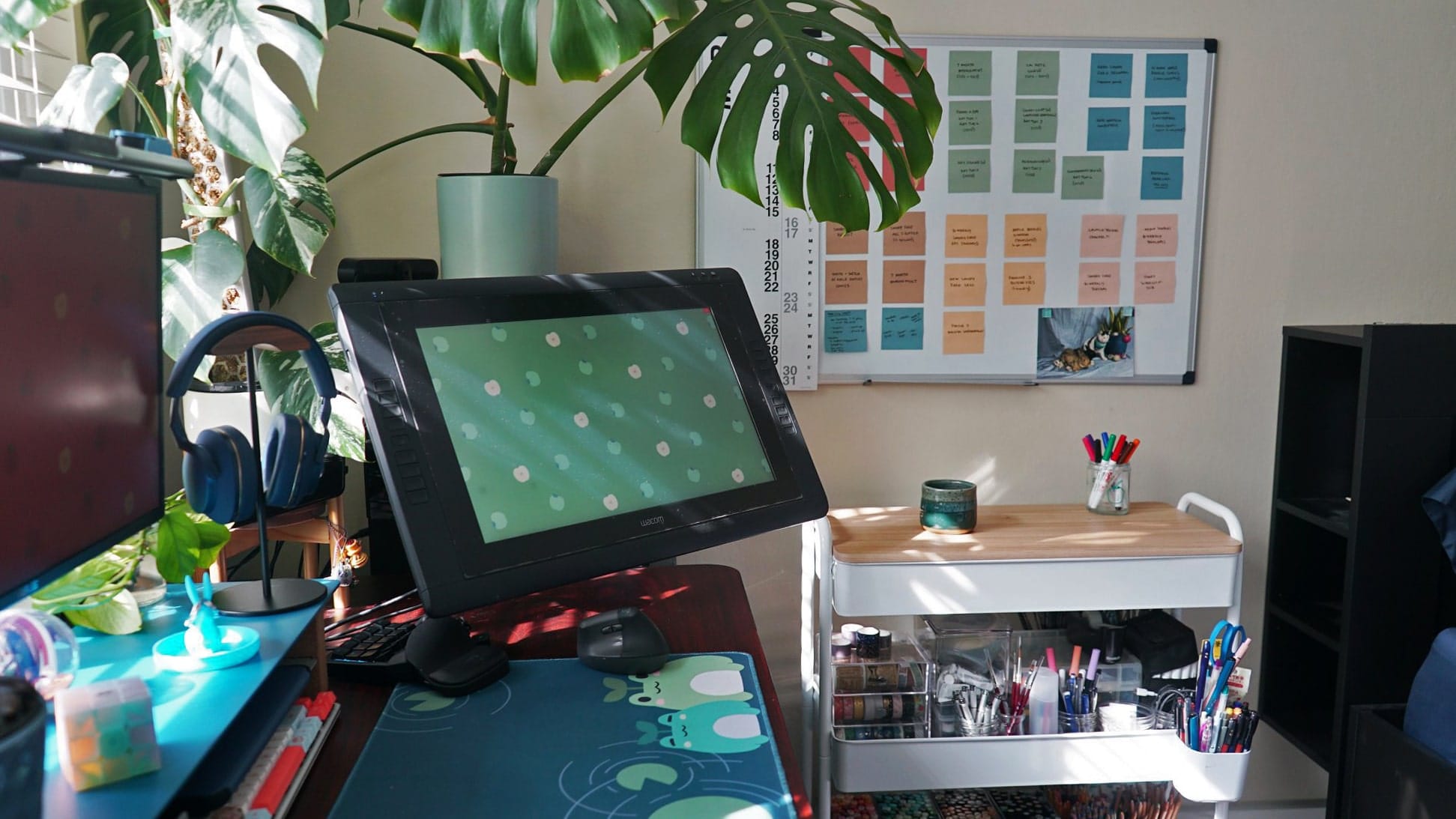A sunlit workspace with a drawing tablet, headphones, indoor plants, and an organised wall planner next to a desk with art supplies
