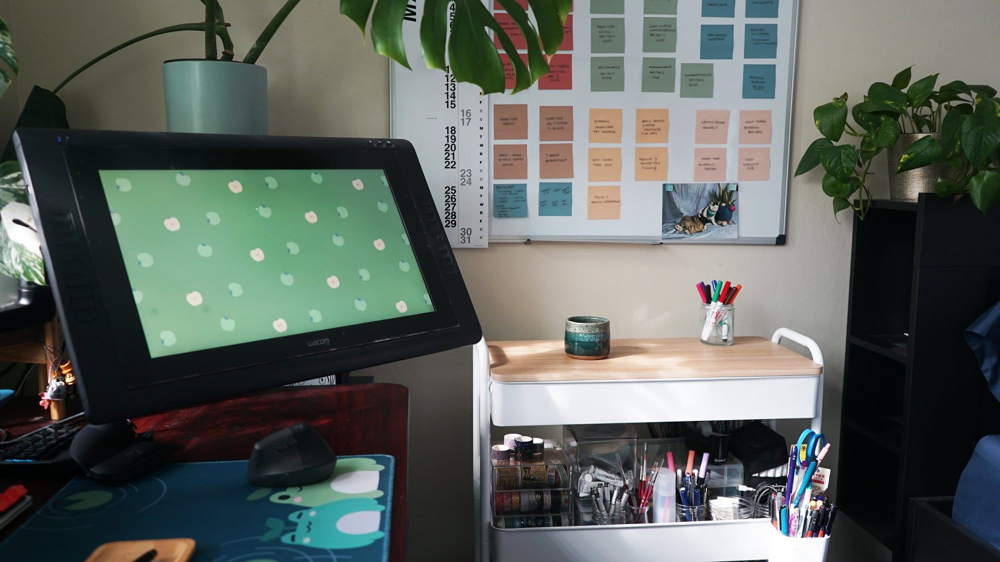 A creative workspace with a digital drawing tablet, an organised wall planner, and a desk with art supplies and potted plants