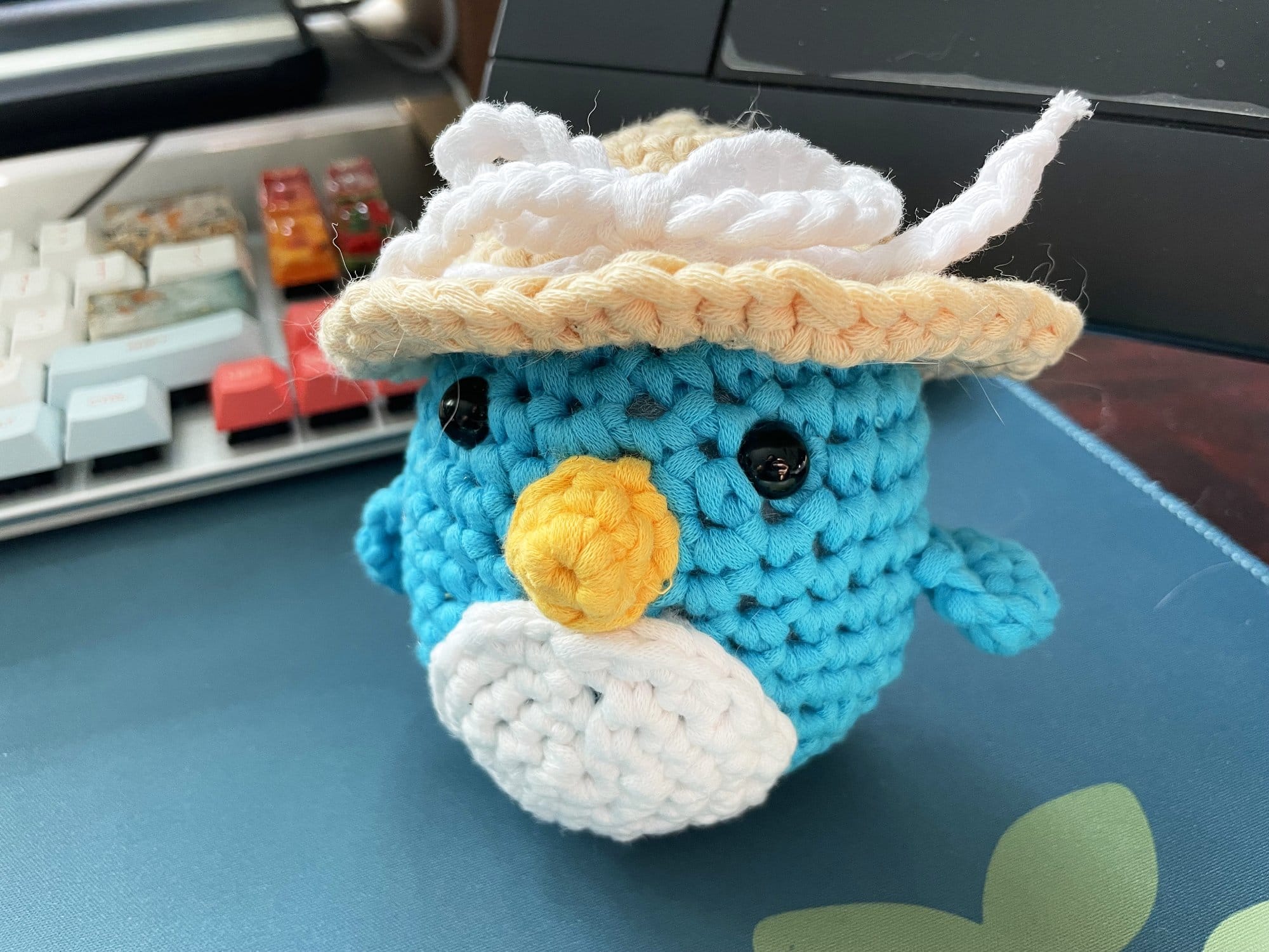 A handcrafted, crochet blue bird wearing a hat, with button eyes and a yellow beak, sitting on a desk