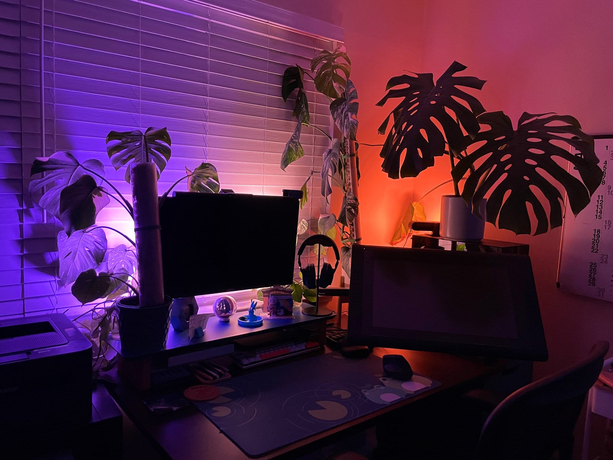 An atmospheric home office illuminated with purple and red hues, featuring a monitor, graphic tablet, and silhouetted plants against the blinds