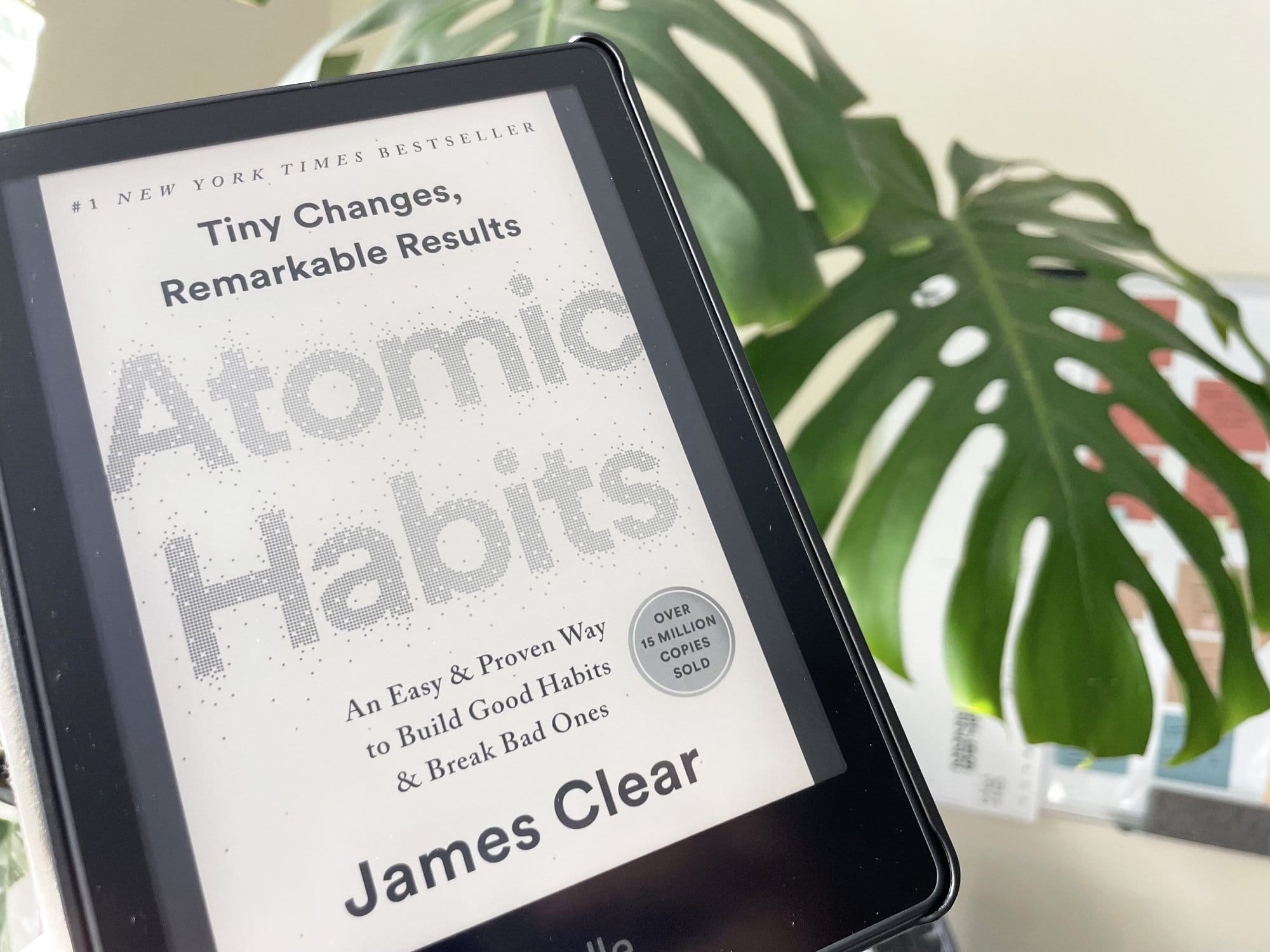An e-reader displaying the cover of “Atomic Habits” by James Clear, with a large monstera leaf in the background