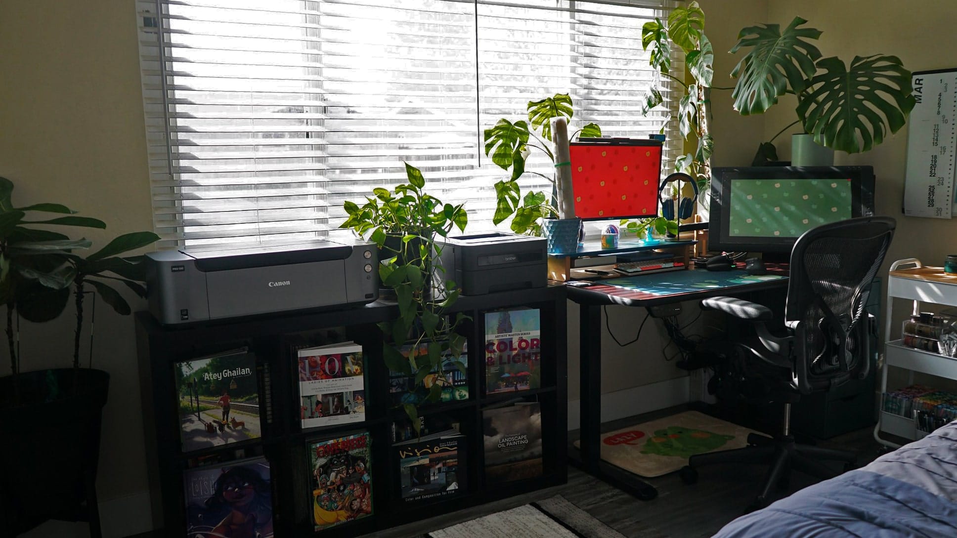 A dimly lit bedroom home office with two printers, a large monitor, indoor plants, and a bookshelf filled with art resources