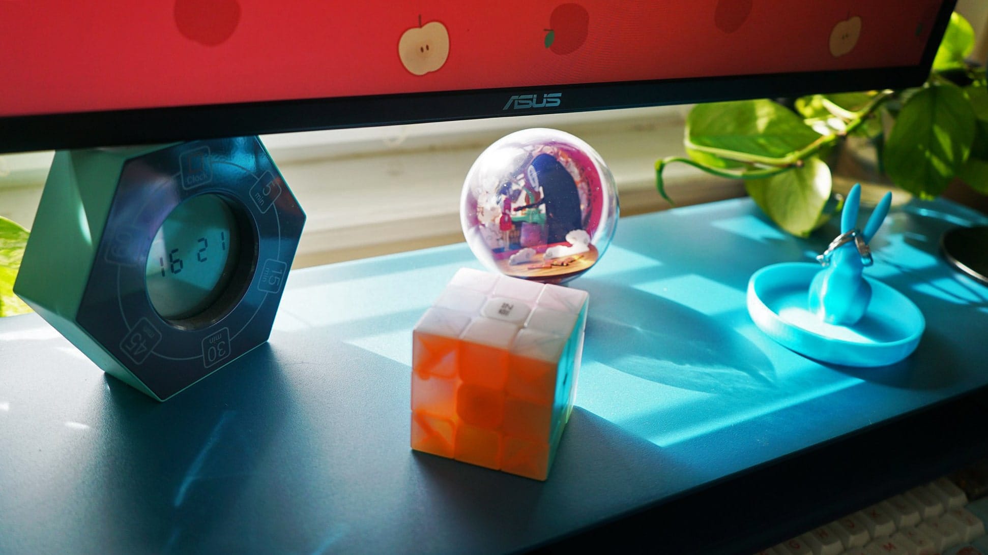 A close-up view of a desk featuring a colourful puzzle cube, a digital clock, a snow globe, and a decorative blue rabbit ring holder