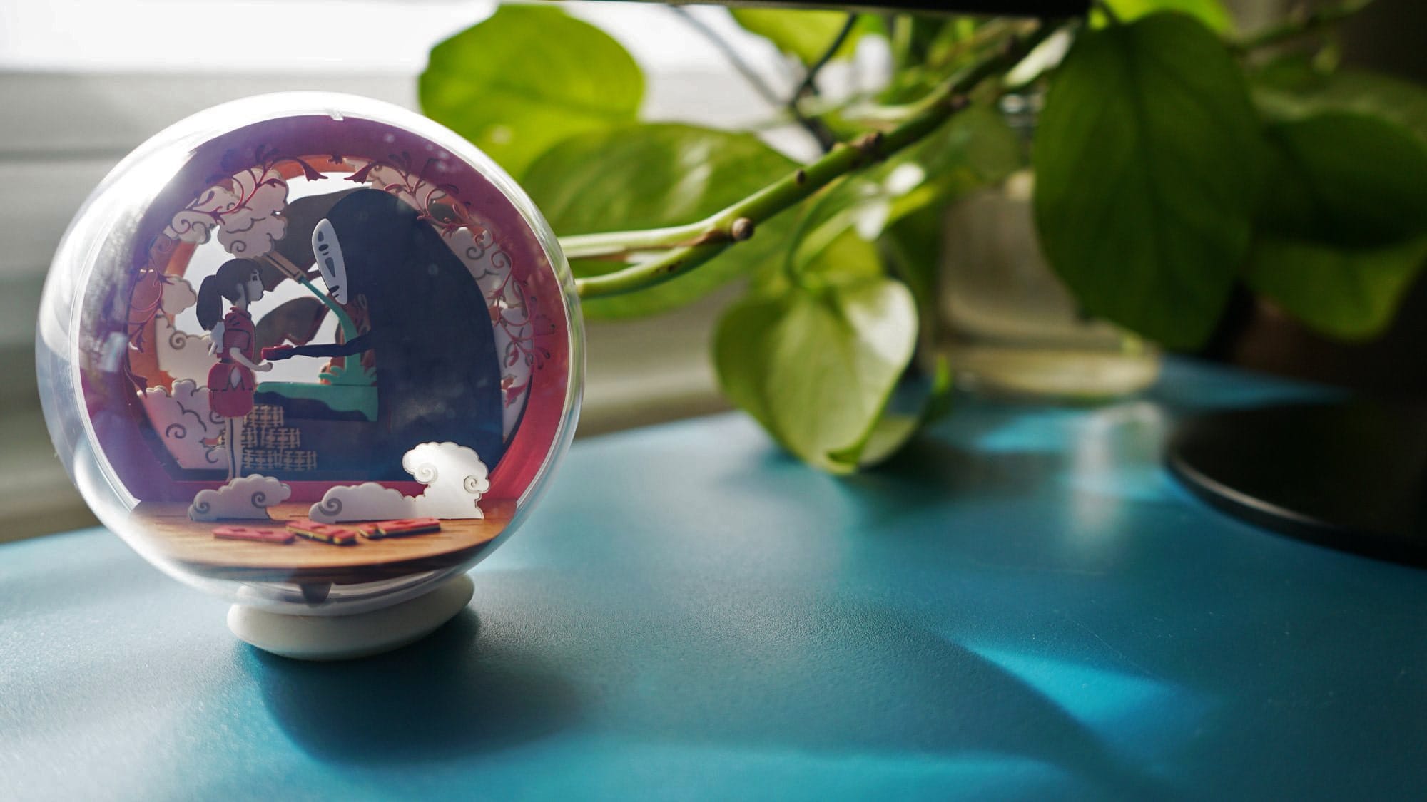 A snow globe on a desk with a scenic depiction inside, next to a green potted plant in soft light