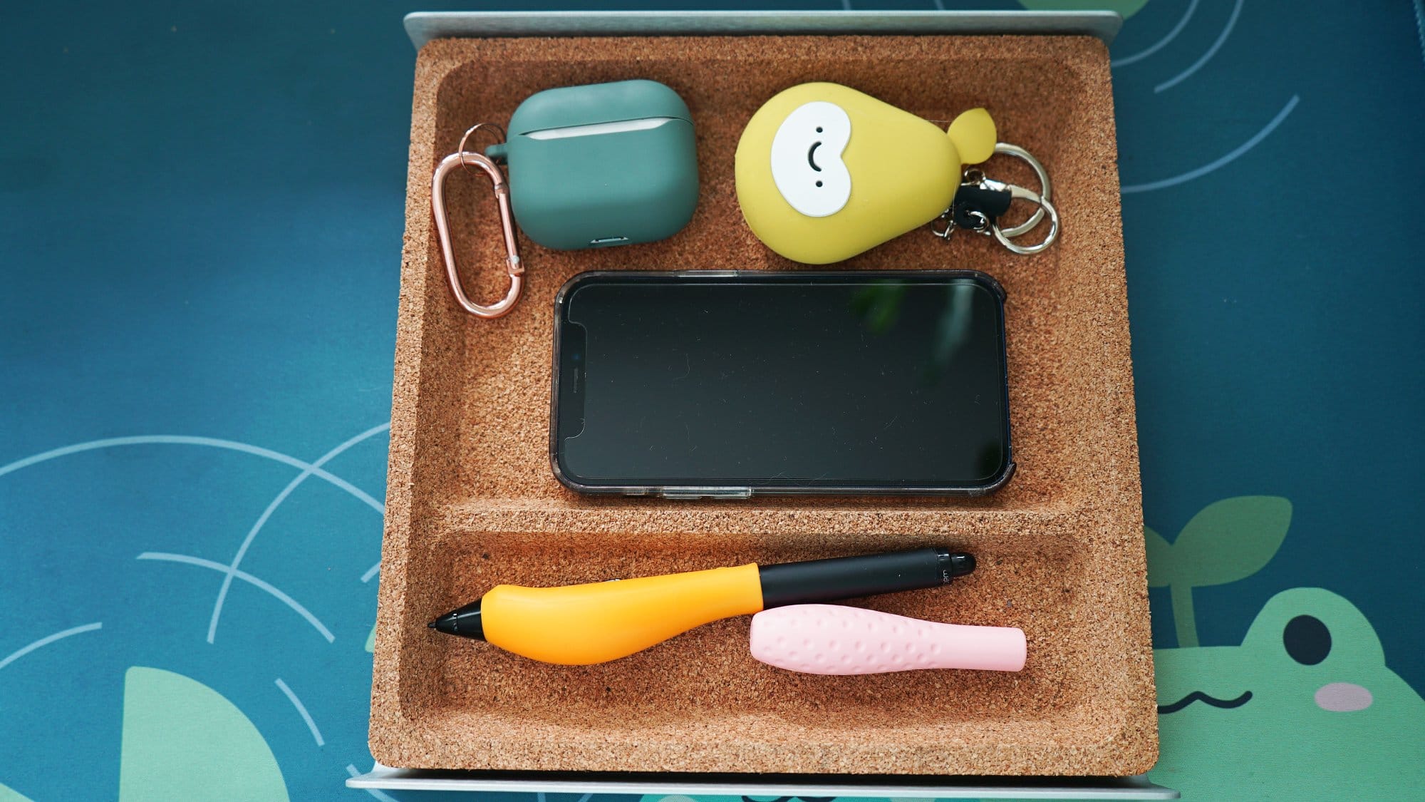  A cork desk organiser holding a smartphone, wireless earbuds case, a keychain with a cute character, and two pens, on a blue desk mat
