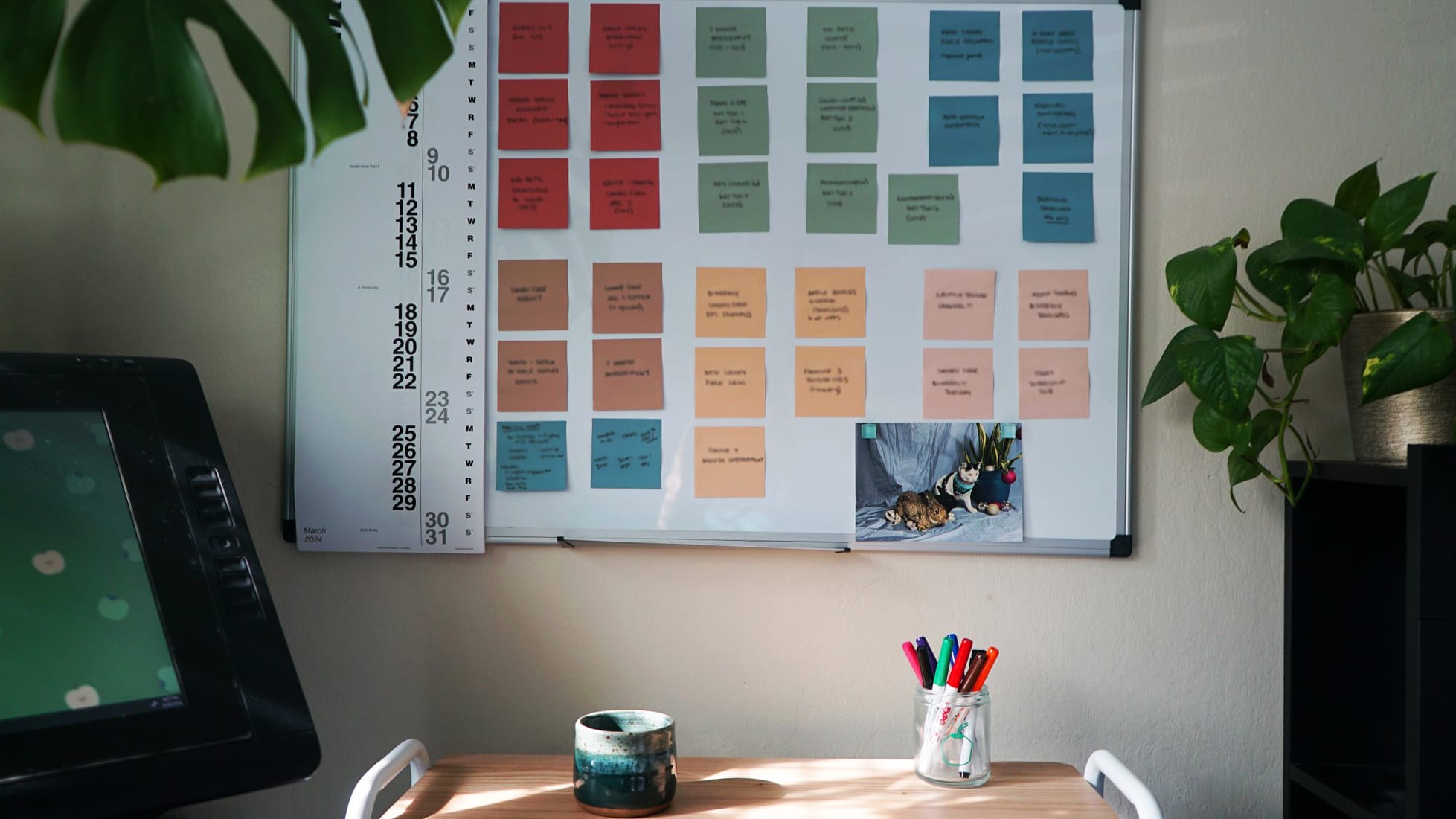 A home office corner with a large wall calendar, a pot of green plants, and a desk with a digital drawing tablet and a jar of colourful markers