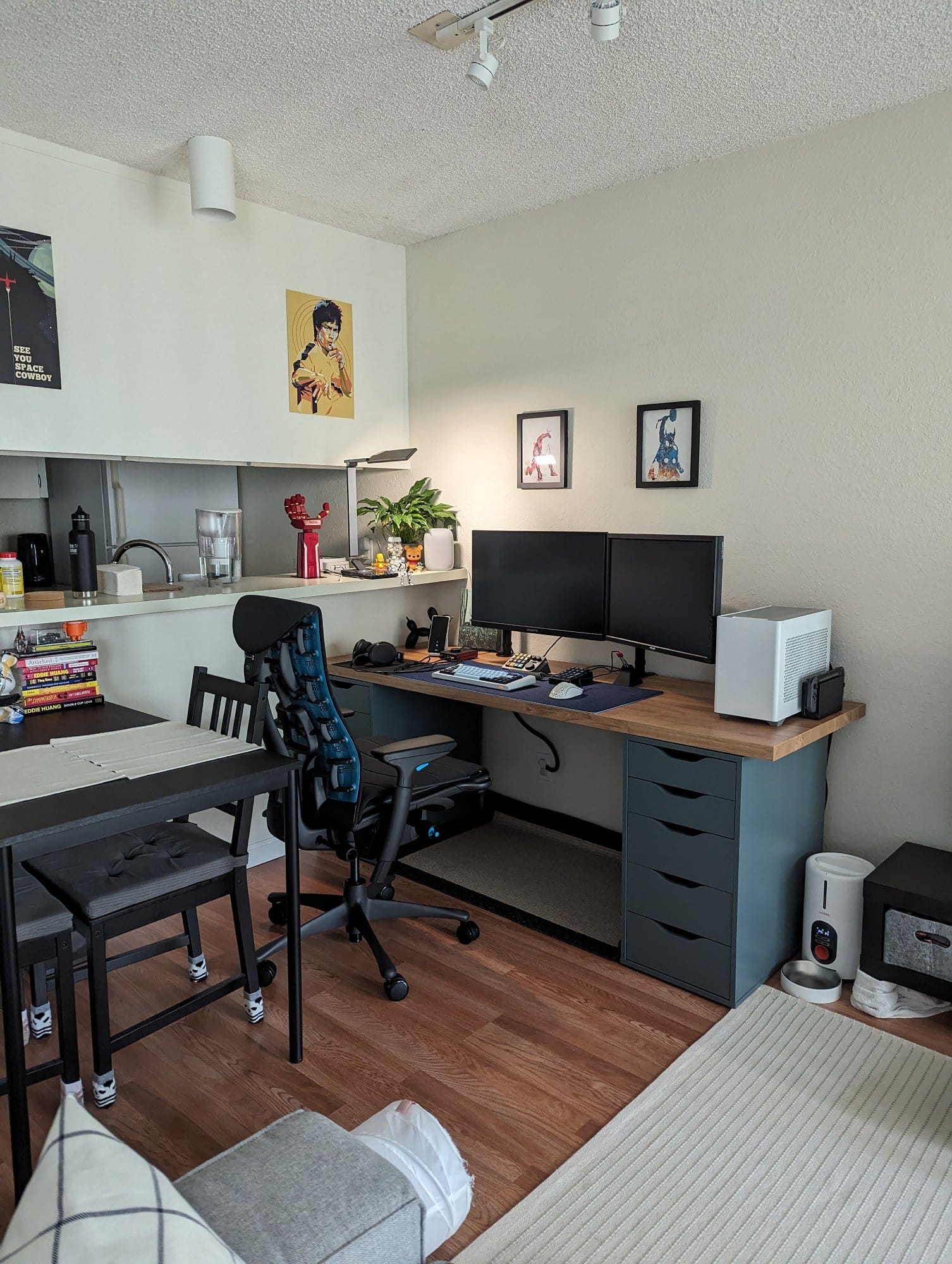 A home office space with an Embody ergonomic chair, a desk with a dual monitor setup, framed artworks on the wall, and a dining area with a black table in the background