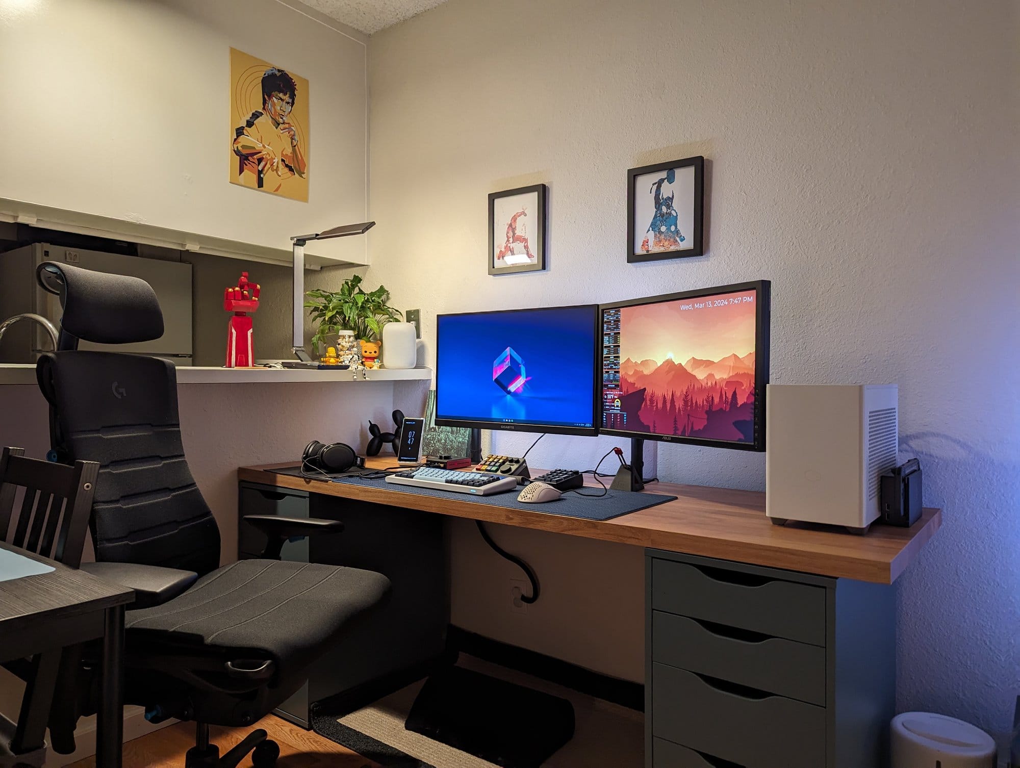 A cosy bedroom home office with an Embody chair, a desk supporting a computer with a lit screen, vibrant framed art on the wall, and decorative items on a shelf above