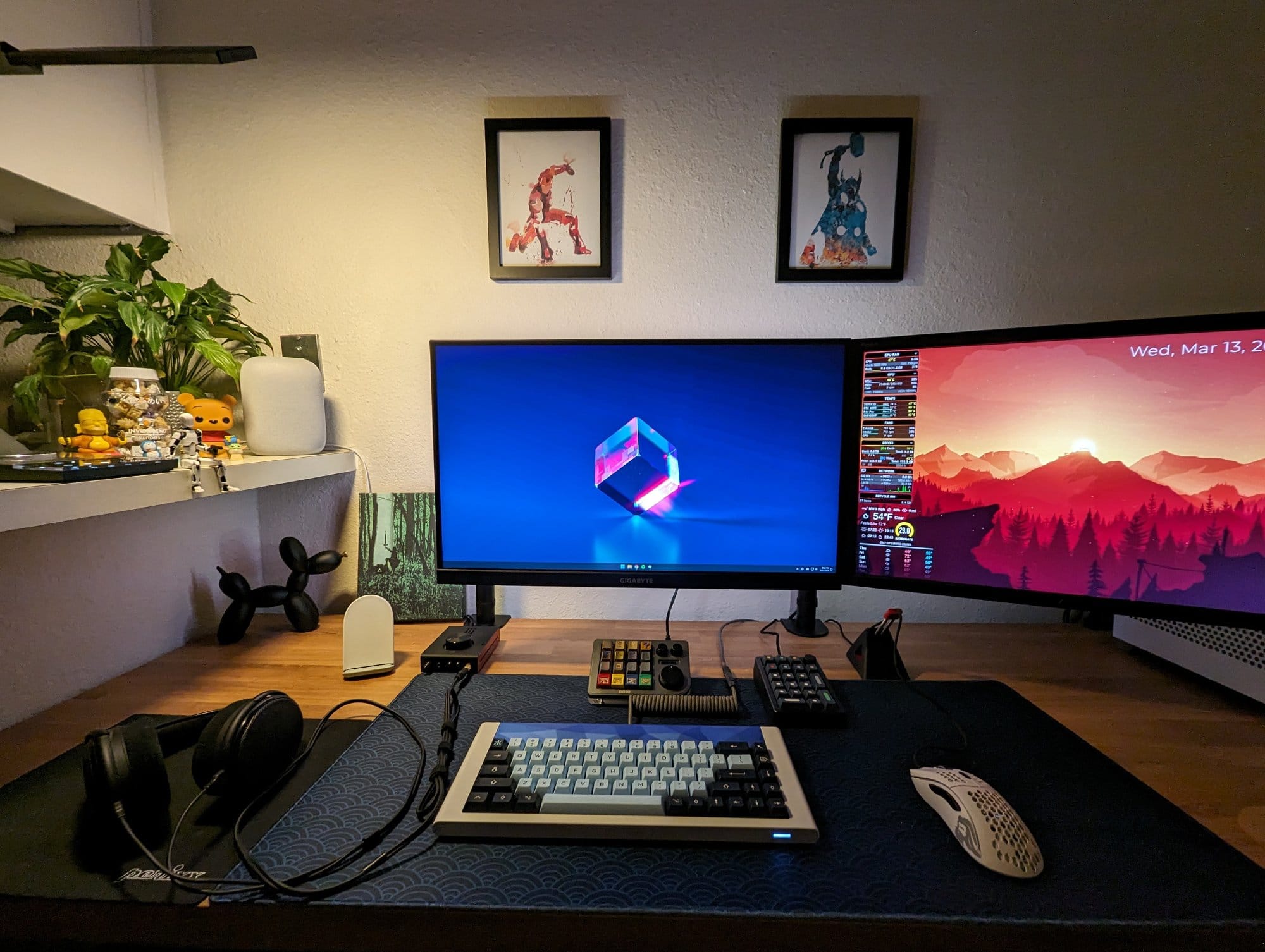 An evening view of a desk with a mechanical keyboard, a pair of headphones, dual monitors, and a collection of figurines and plants on the shelf above
