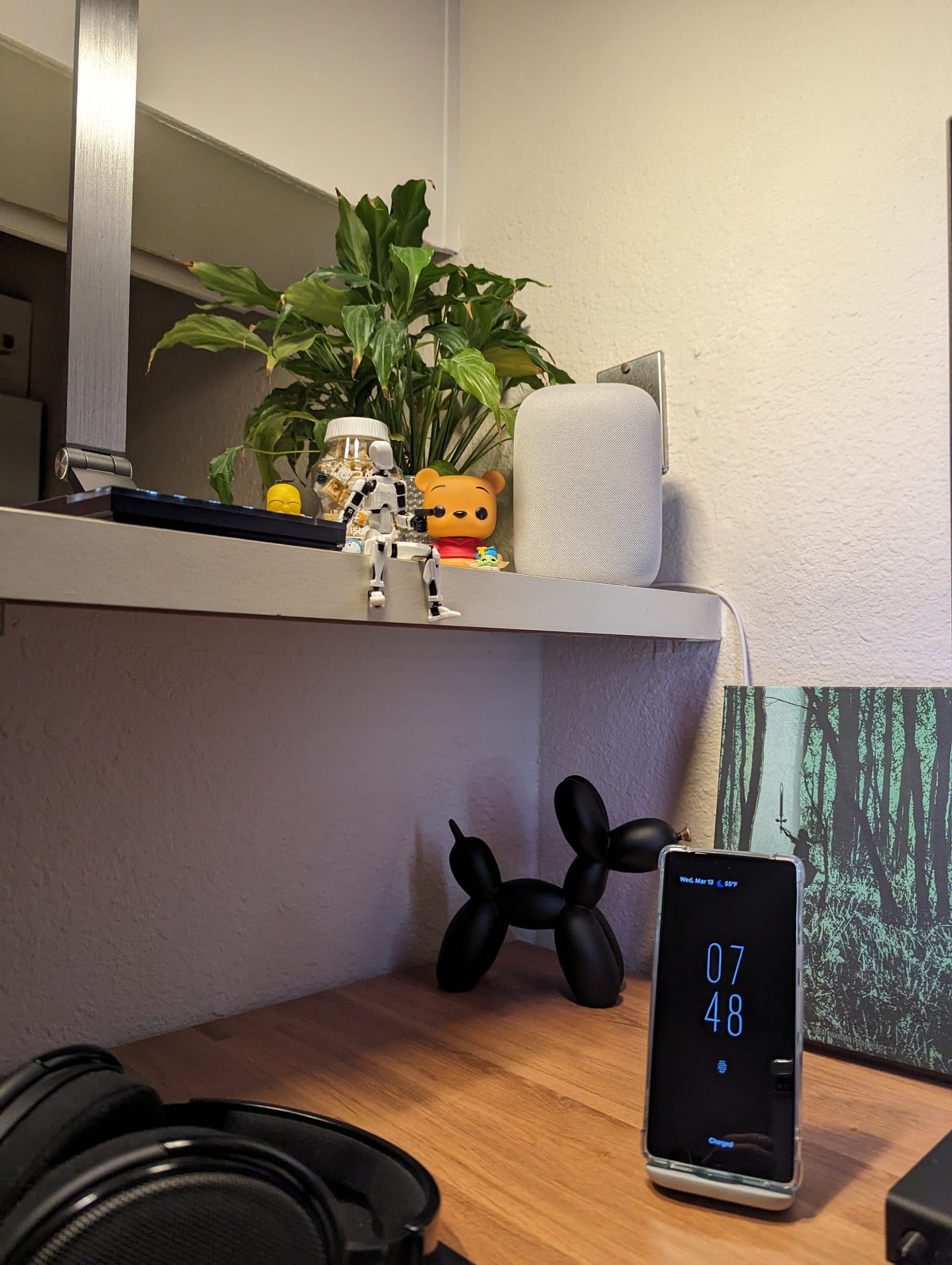 A shelf with various items including a plant, collectible figures, a speaker, and a smartphone displaying the time against a backdrop of a framed forest picture