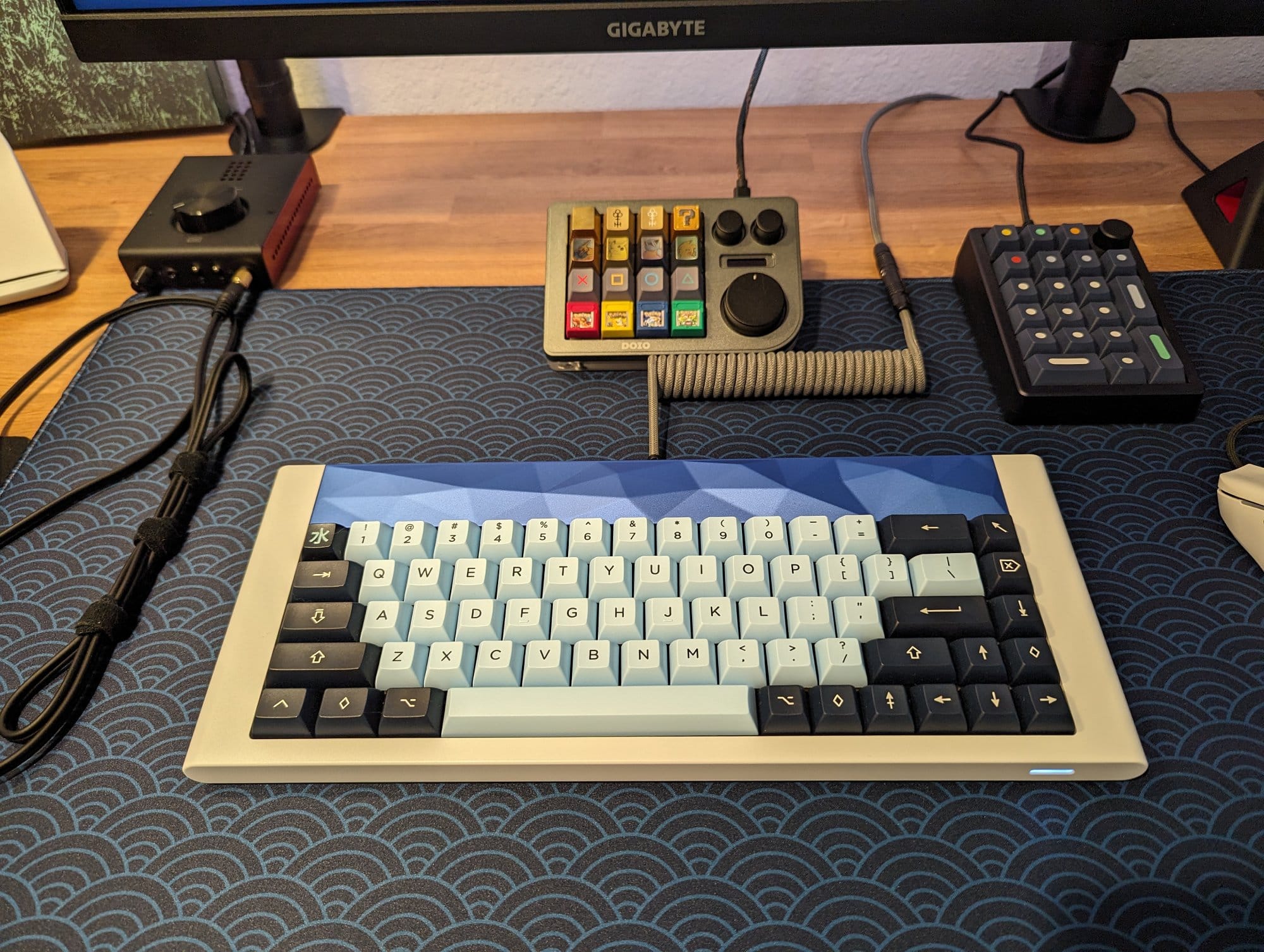 A close-up of a desk with a custom mechanical keyboard in blue and grey, surrounded by various audio equipment and a computer monitor in the background