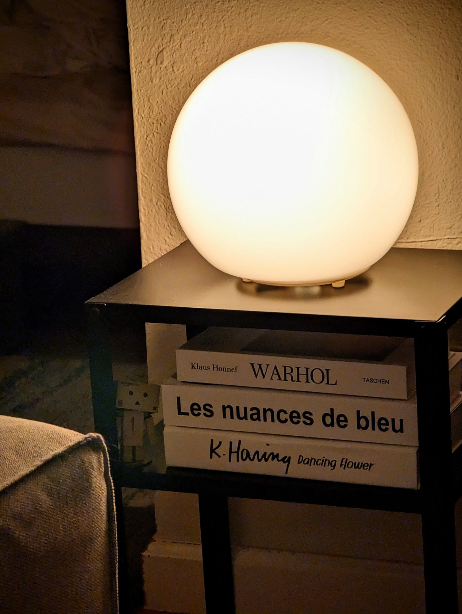 A softly glowing spherical lamp atop a black side table with art books and a small cardboard figure nestled below