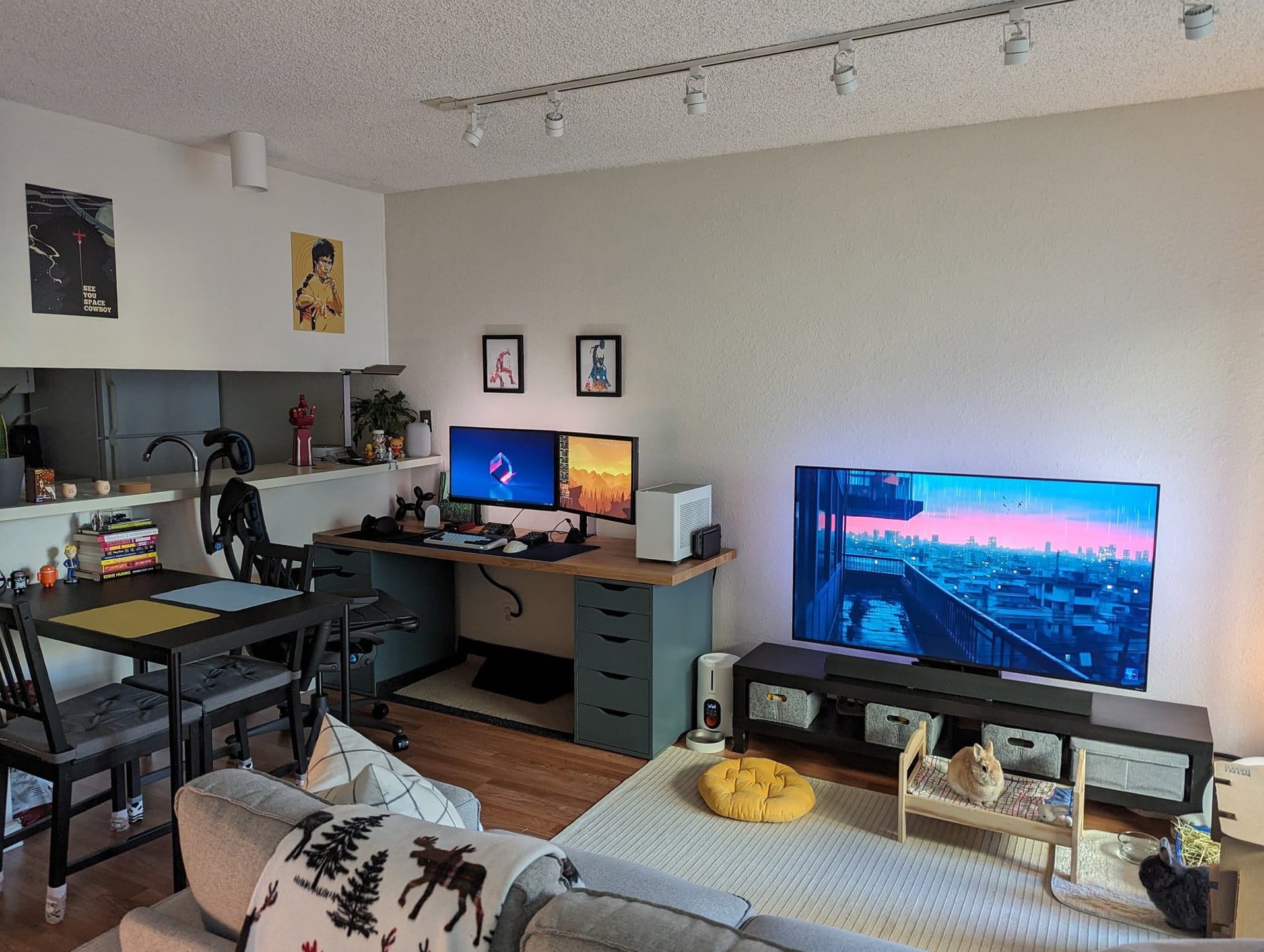 A cosy living space with a large TV opposite the sofa, a dining area to the left, and a home office setup with dual monitors, and artworks on the wall
