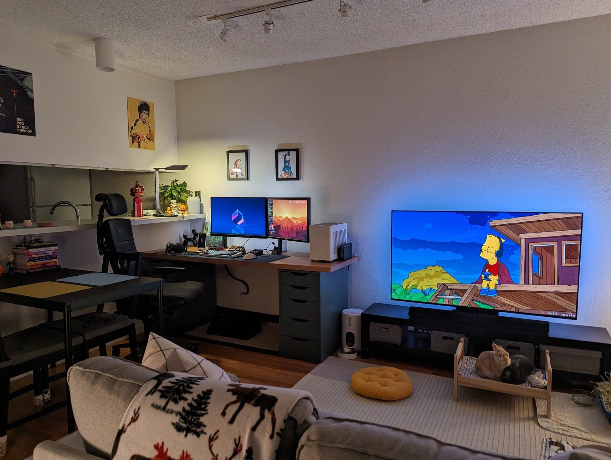 A living room featuring a large TV showing the Simpsons series, a comfortable sofa with a patterned throw, and an office area with dual monitors and desk in the corner