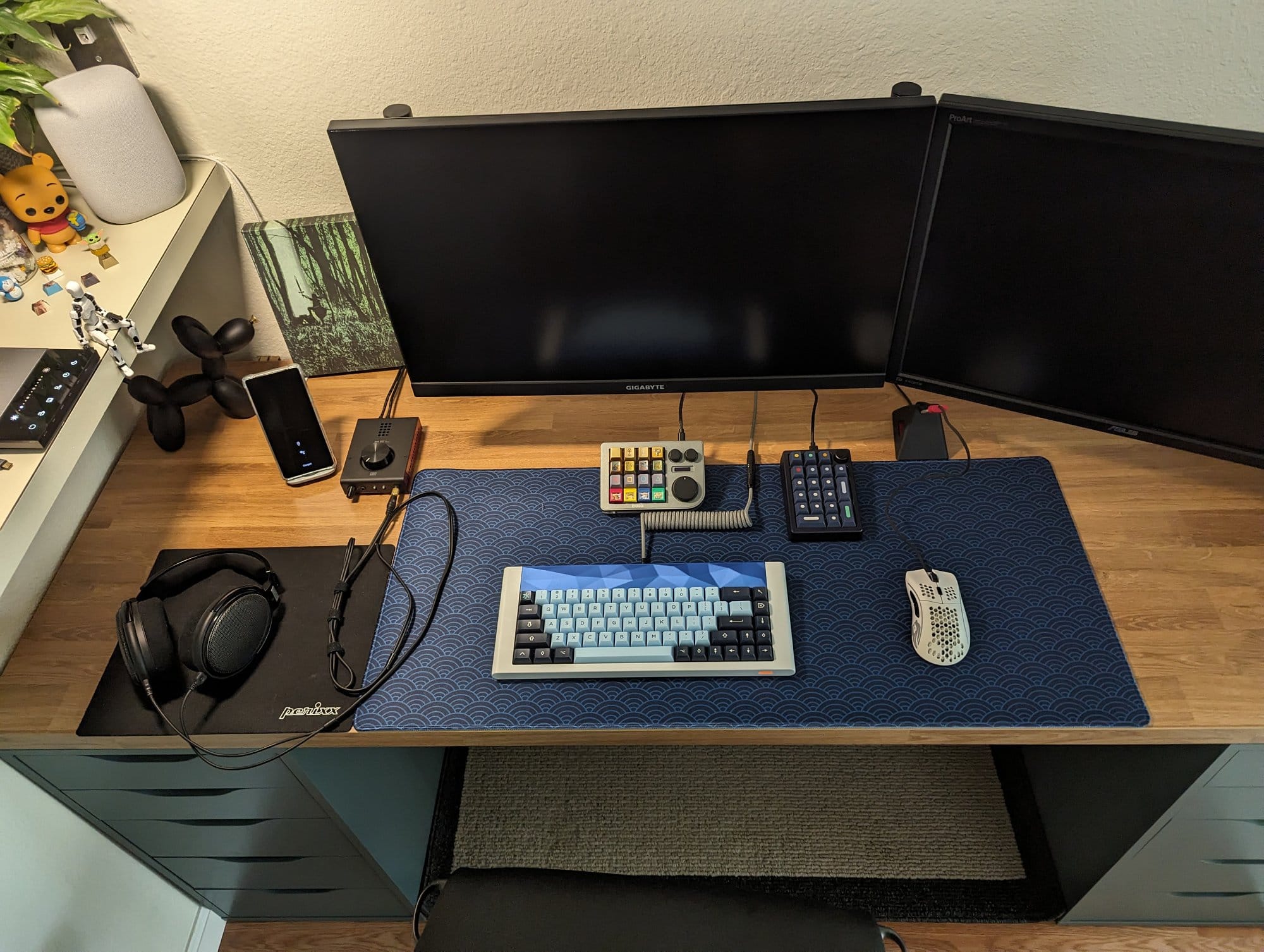 A neat desk setup with two turned-off monitors, a mechanical keyboard, headphones on a mat, and various desk accessories, with a shelf holding small figurines to the side
