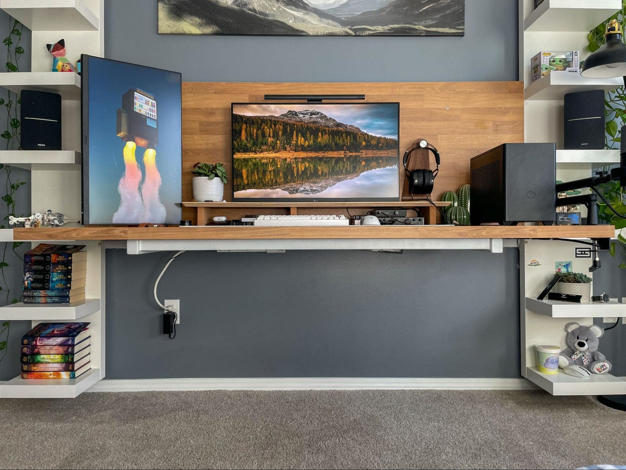 A tidy workstation with a wooden desk and shelves, featuring dual monitors, speakers, and decorative items, set against a grey wall with mountainous artwork