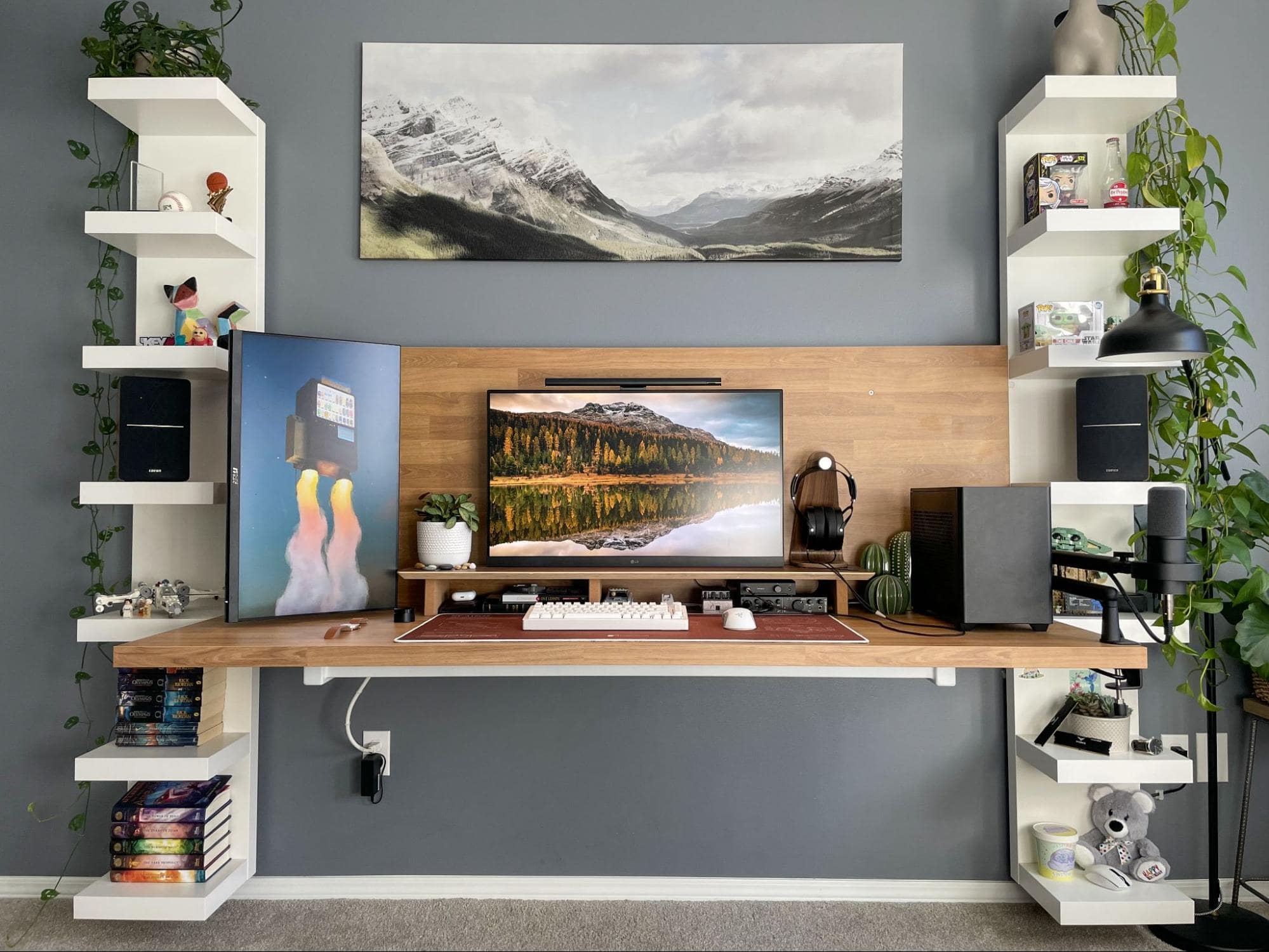 A modern home office setup with a floating desk flanked by white shelves filled with books and decor, two monitors, and framed artwork against a grey wall, complemented by indoor plants