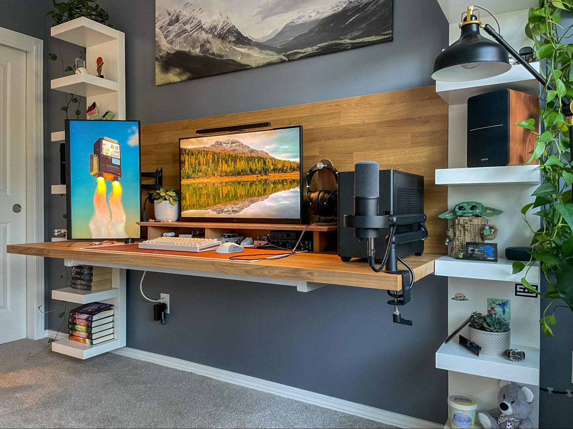 A well-organised home office with a floating wooden desk, dual monitors, a microphone, speakers, and decorative shelves with plants and toys, set against a grey wall with landscape paintings