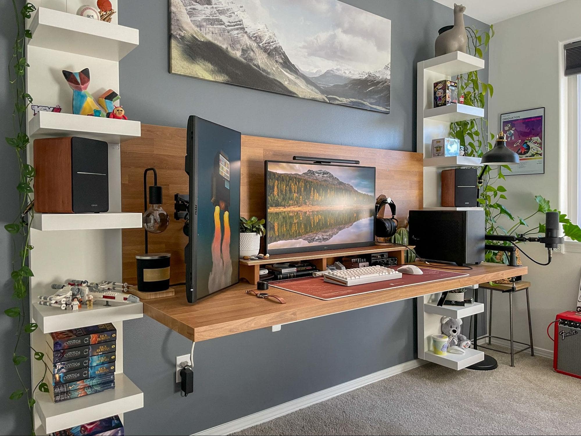 A stylish home office featuring a large wooden desk with dual monitors, a microphone, a mechanical keyboard, and a pair of headphones, flanked by white shelves with playful decor, all set against a grey wall with mountain canvas art