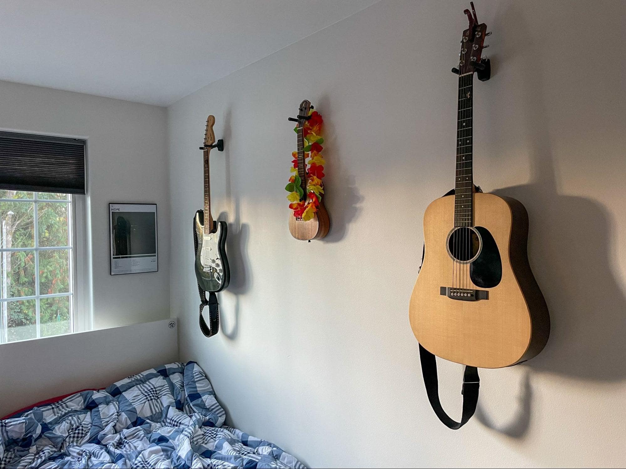Guitars hung on a white wall above a bed with blue and white bedding, next to a window with a view of greenery outside
