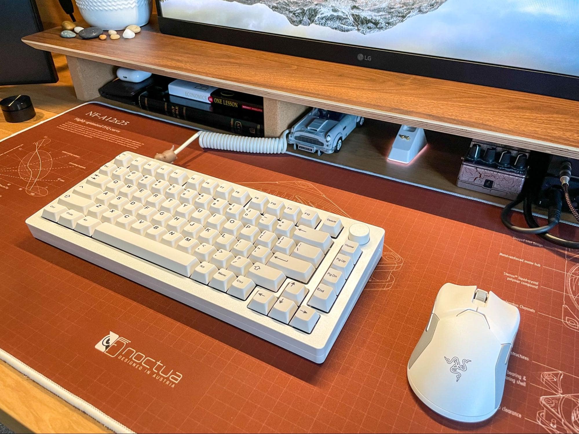 A close-up of a desk with a mechanical keyboard, white gaming mouse, and a large monitor, with a schematic mouse pad and tech accessories in the background