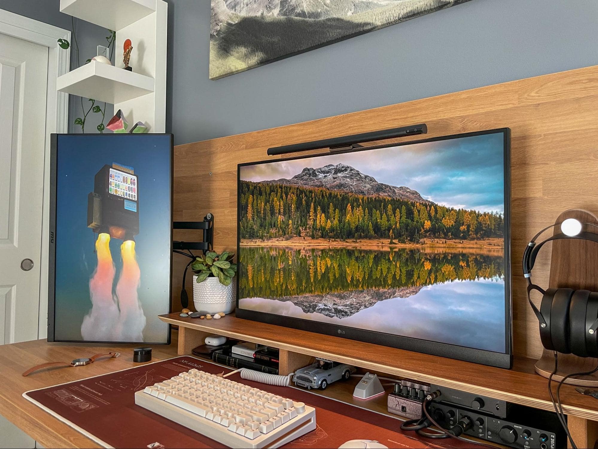A detailed view of a home office featuring a large monitor, mechanical keyboard, and decorative items on a desk, with a scenic mountain landscape on the screen and grey walls adorned with shelves and artwork in the background