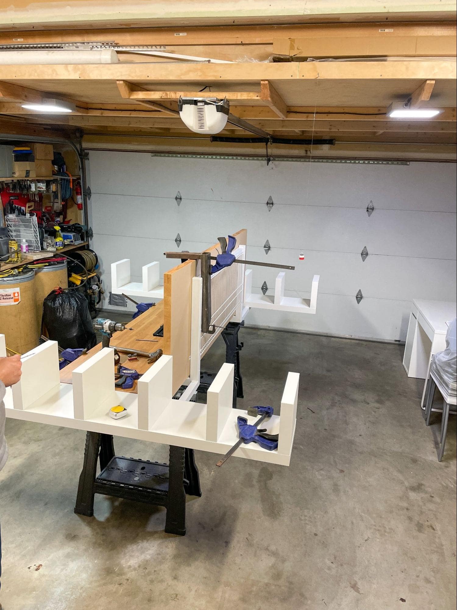 A garage workshop with a workbench holding a wooden construction project, surrounded by various tools and equipment, under exposed wooden ceiling beams