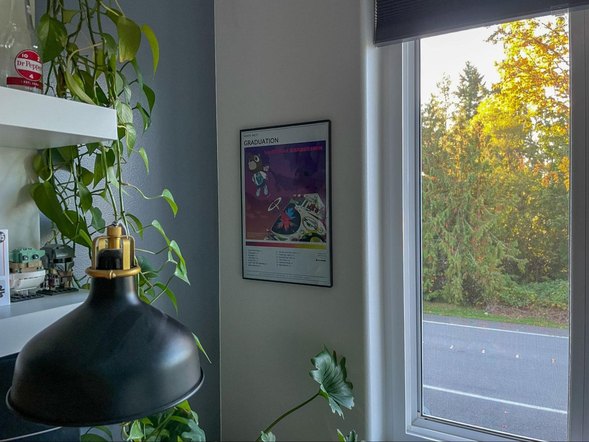 A corner of a room showing a framed poster on a grey wall, a desk lamp, and a window with a view of trees with green foliage
