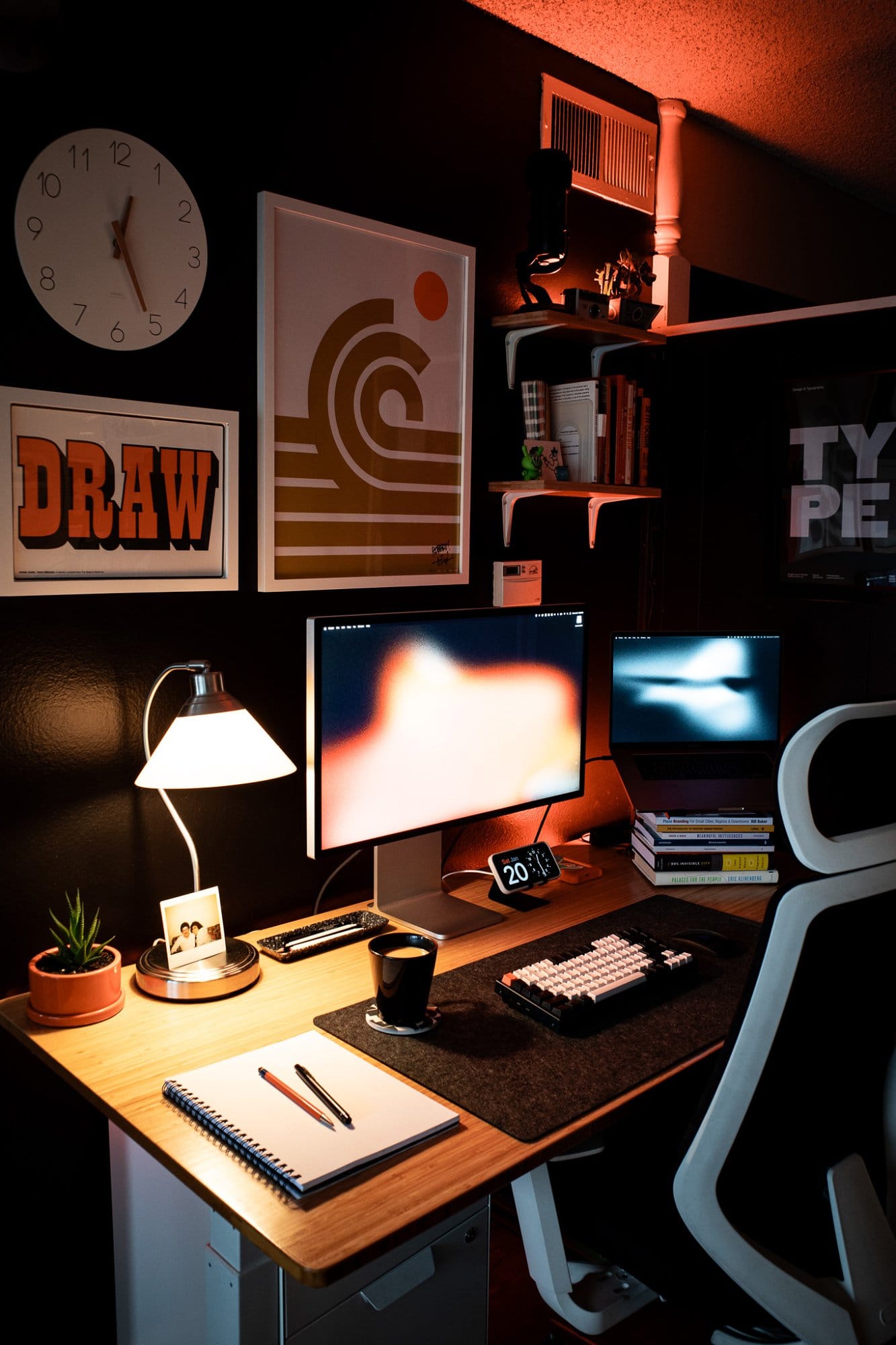 A modern workspace with a wall clock, art prints, shelves, a desk lamp, a mug, a plant, and stationery, with an office chair to the side
