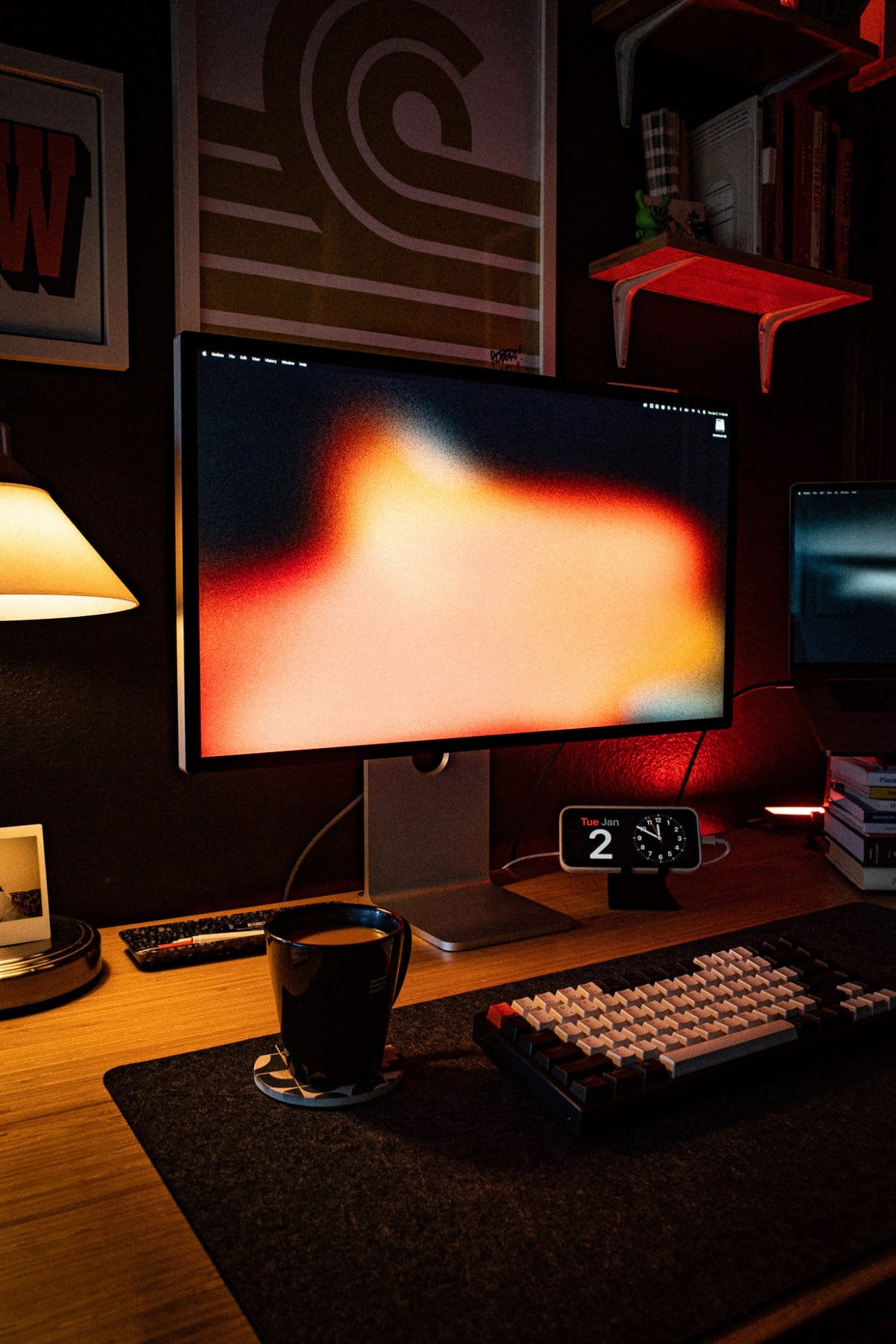 A neat desk with a computer monitor, a cup, and a keyboard under a table lamp, with art and shelves on the wall
