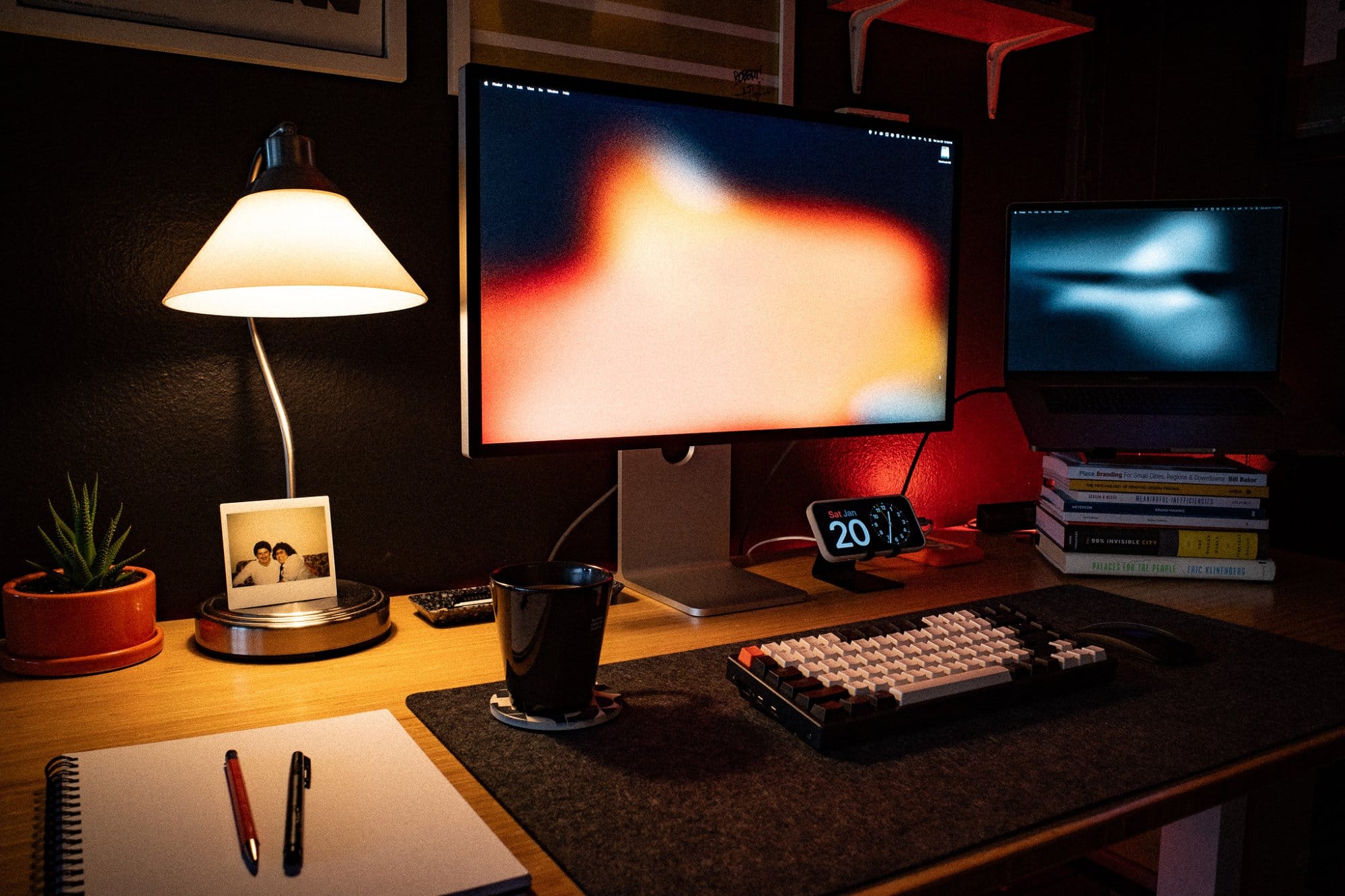 A cosy and inviting desk setup with a lamp, an Apple Studio Display, a MacBook, a mechanical keyboard, and a mug, complemented by a plant and personal photo