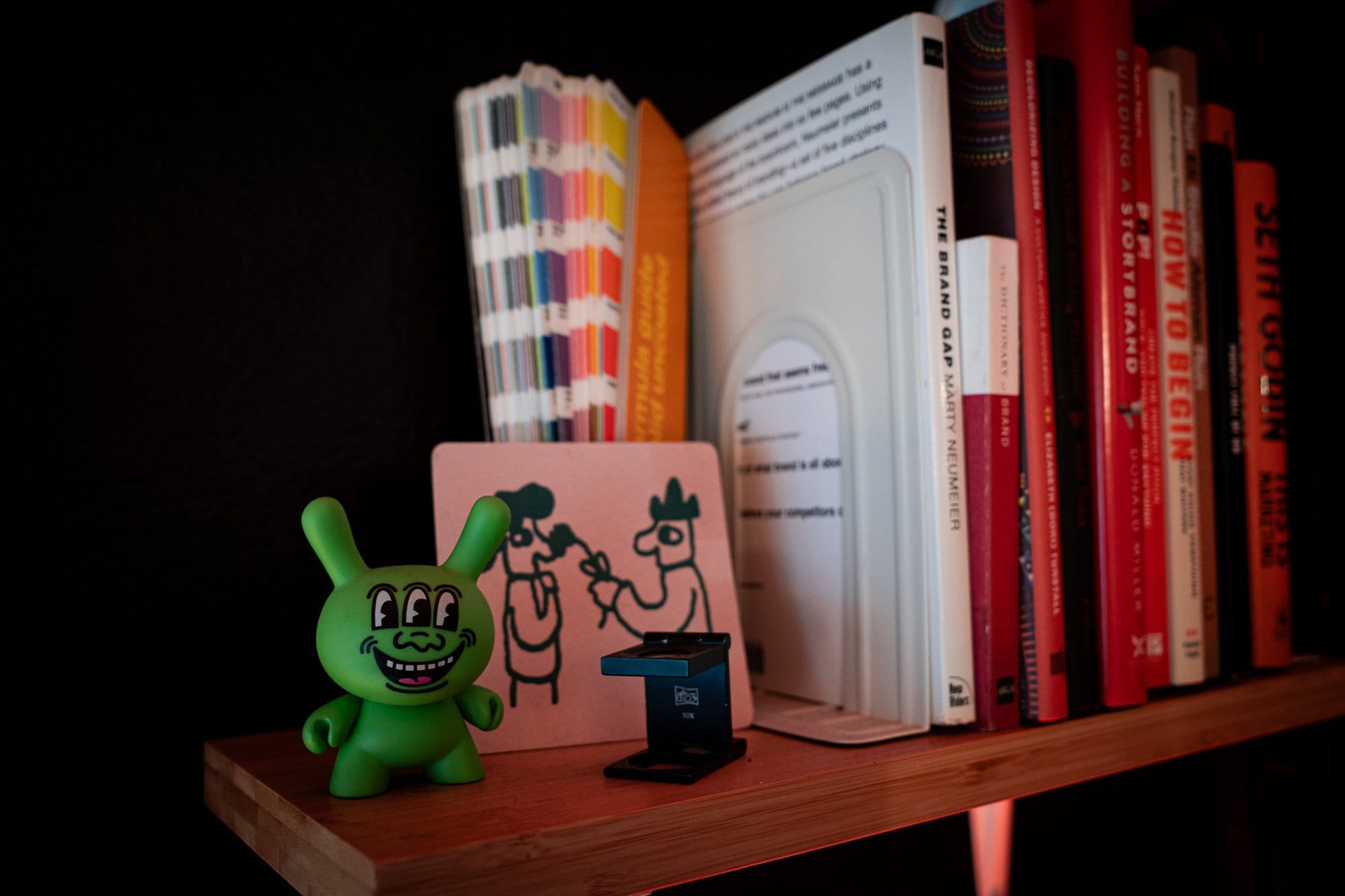 A shelf with a colourful array of books and a whimsical Keith Haring’s Three Eyed Face 8″ Dunny art figure, against a dark background