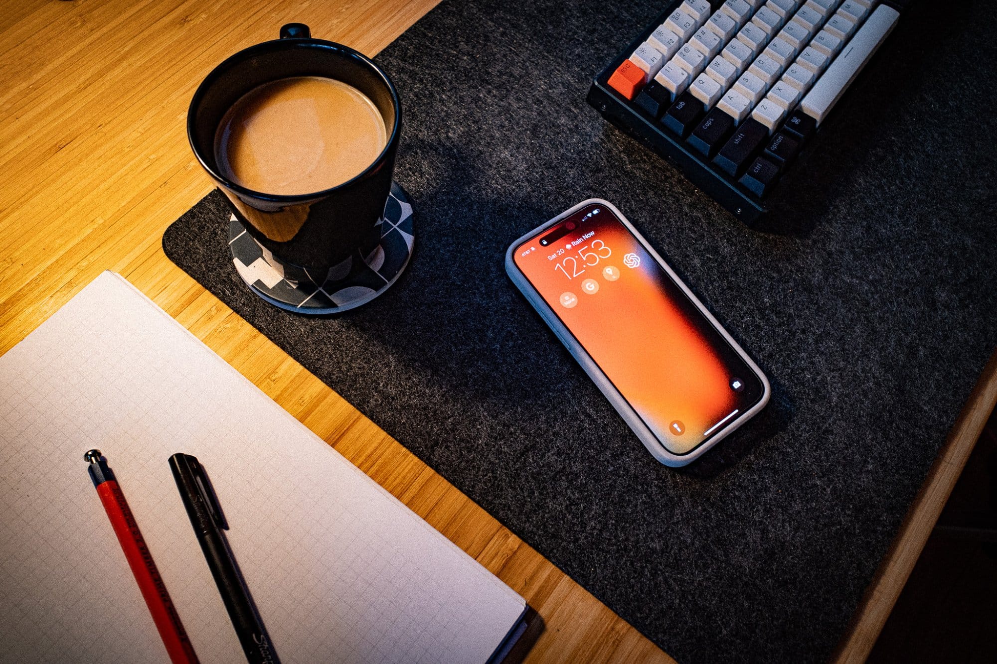 A mug of coffee, a mobile phone displaying the time, a notepad with pens, and a mechanical keyboard on a desk mat