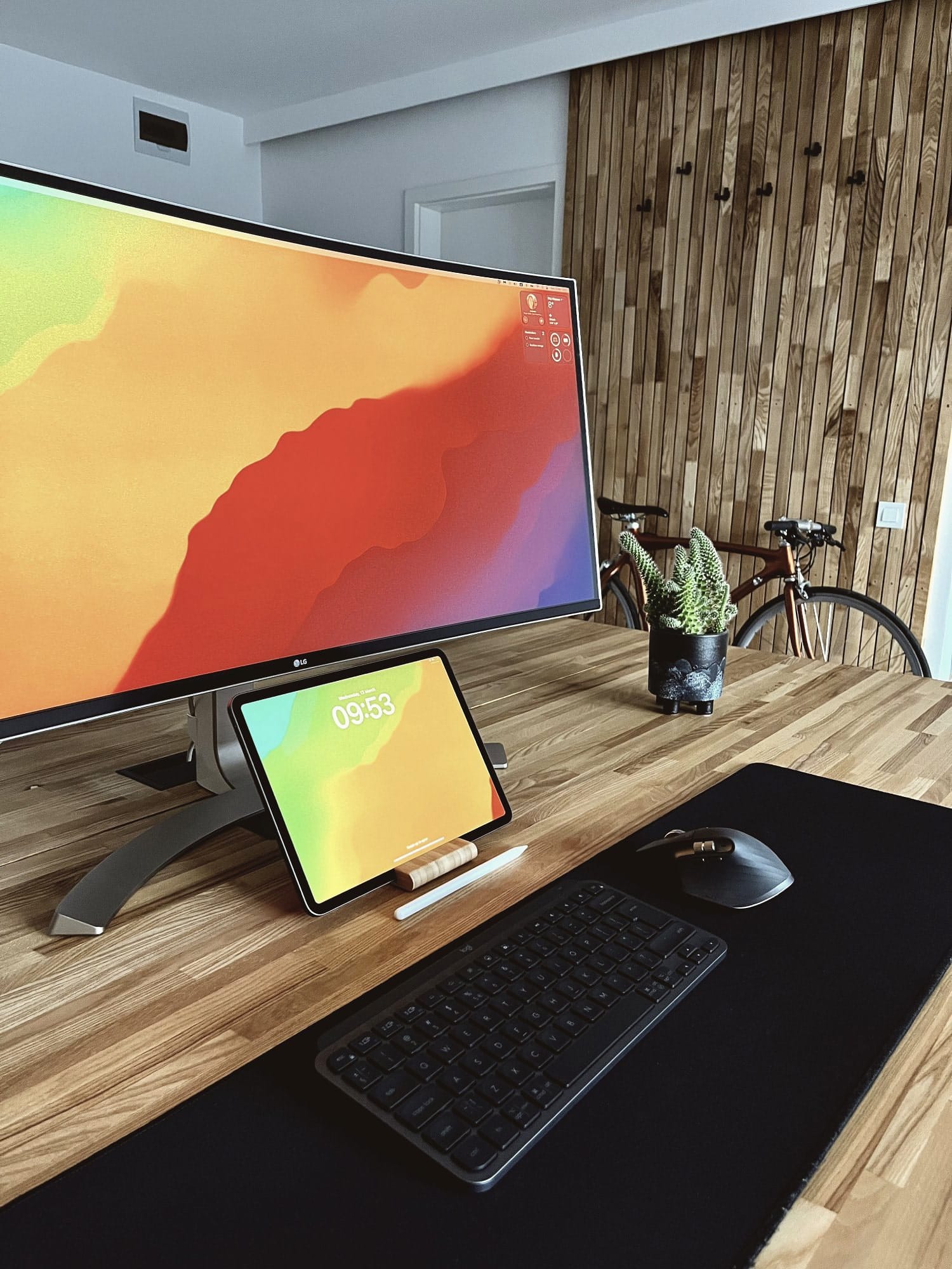 A stylish workspace setup featuring a widescreen monitor, a wireless keyboard, and a tablet on a wooden desk, with a decorative wooden wall and a bicycle in the background