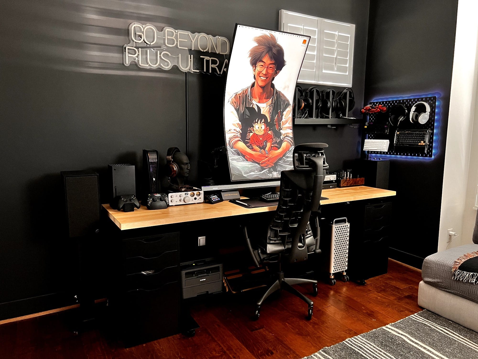 A sleek gaming setup featuring a large curved monitor, an ergonomic Herman Miller Embody chair, audio equipment, and gaming consoles in a minimalist room with a grey sofa in the corner
