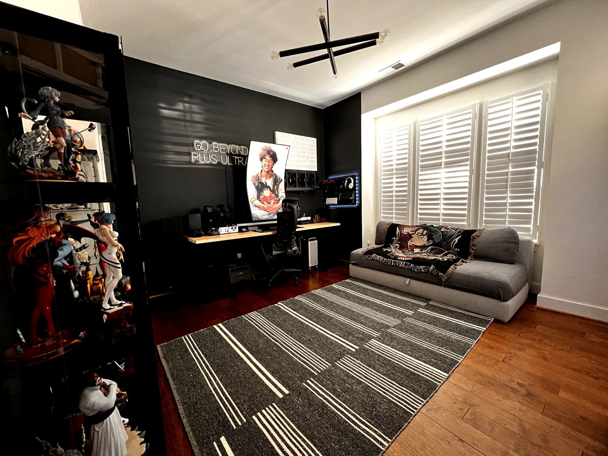 A spacious and stylish room featuring a gaming setup with a large curved monitor, a “GO BEYOND PLUS ULTRA” LED sign, a black ergonomic chair, and shelves displaying action figures