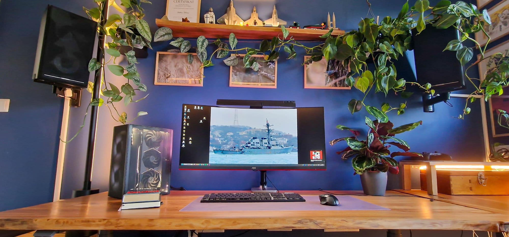 A serene and stylish home office setup featuring a wooden desk with a gaming monitor, surrounded by lush indoor plants, decorative items on a shelf above, and ambient lighting