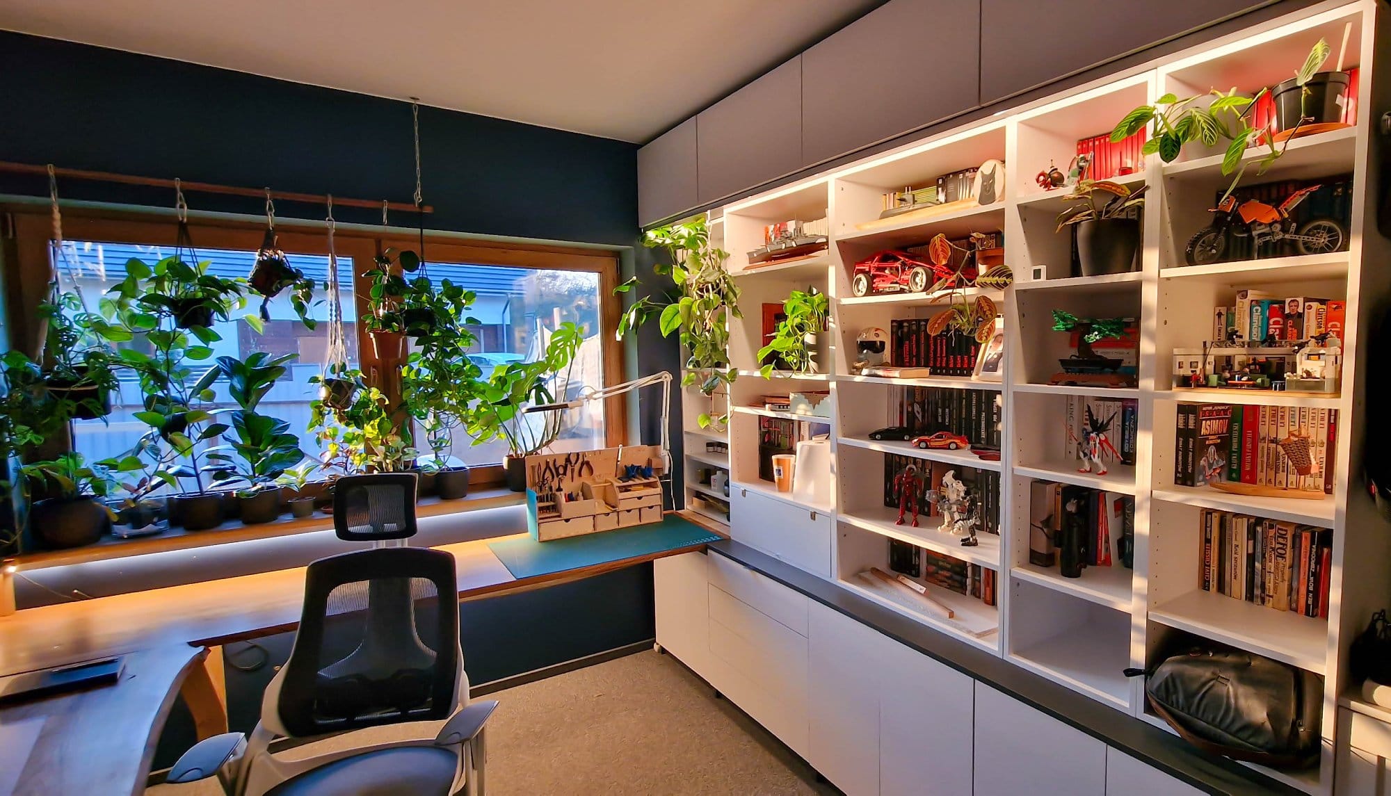 A modern home office with large windows draped with green plants, an ergonomic chair facing a desk with crafting tools, and extensive white shelving filled with books, models, and collectibles