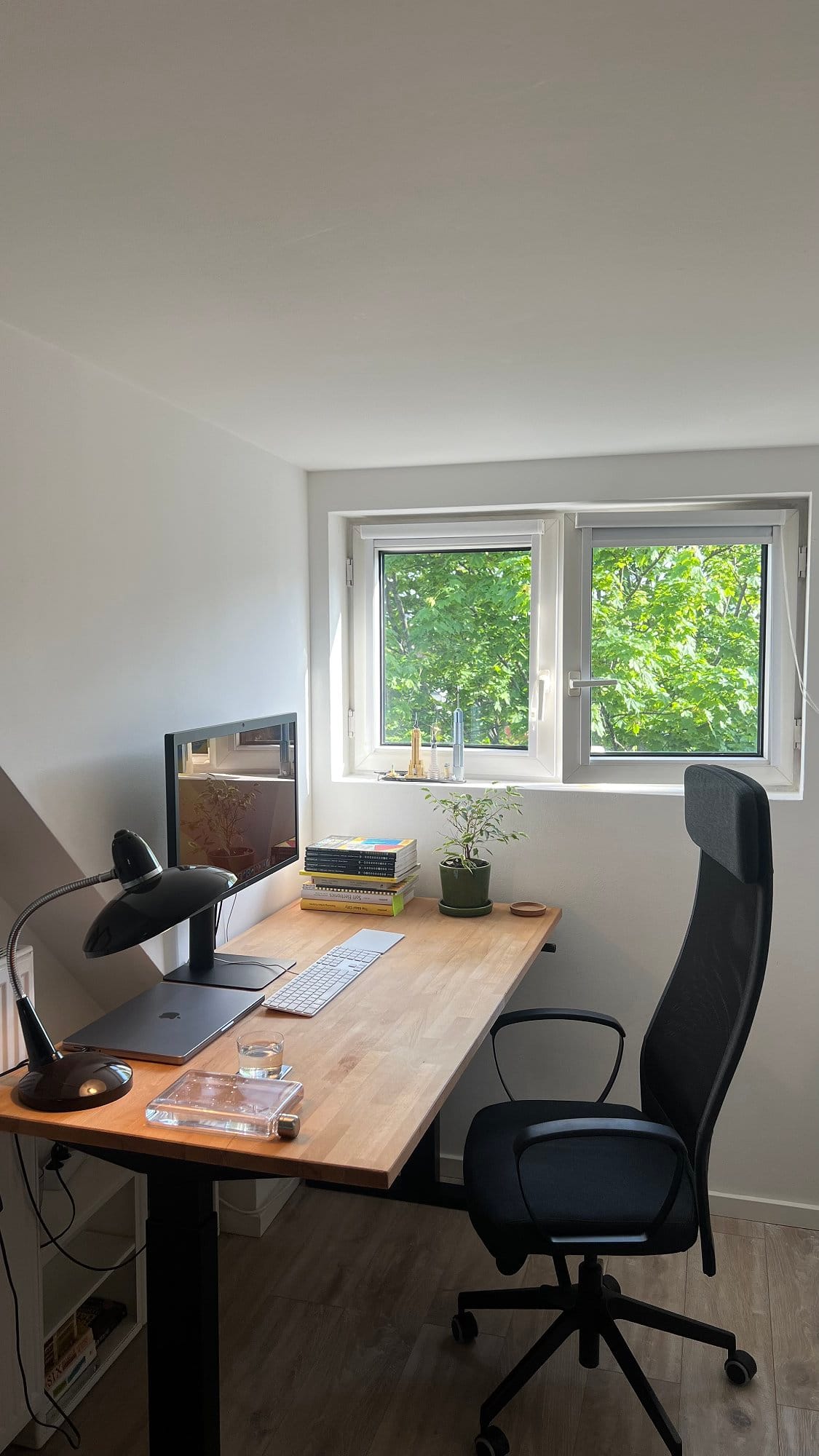 A minimalist home office setup with an LG UltraFine 5K monitor, MacBook Pro M1 16″ 2021, Apple Magic Keyboard, Apple Trackpad II, black desk lamp, clear water bottle, and a glass of water, all arranged on an IKEA GERTON tabletop with a Flexispot H2 standing desk frame, IKEA MARKUS chair, a stack of books, a small plant, and a LEGO model on the windowsill