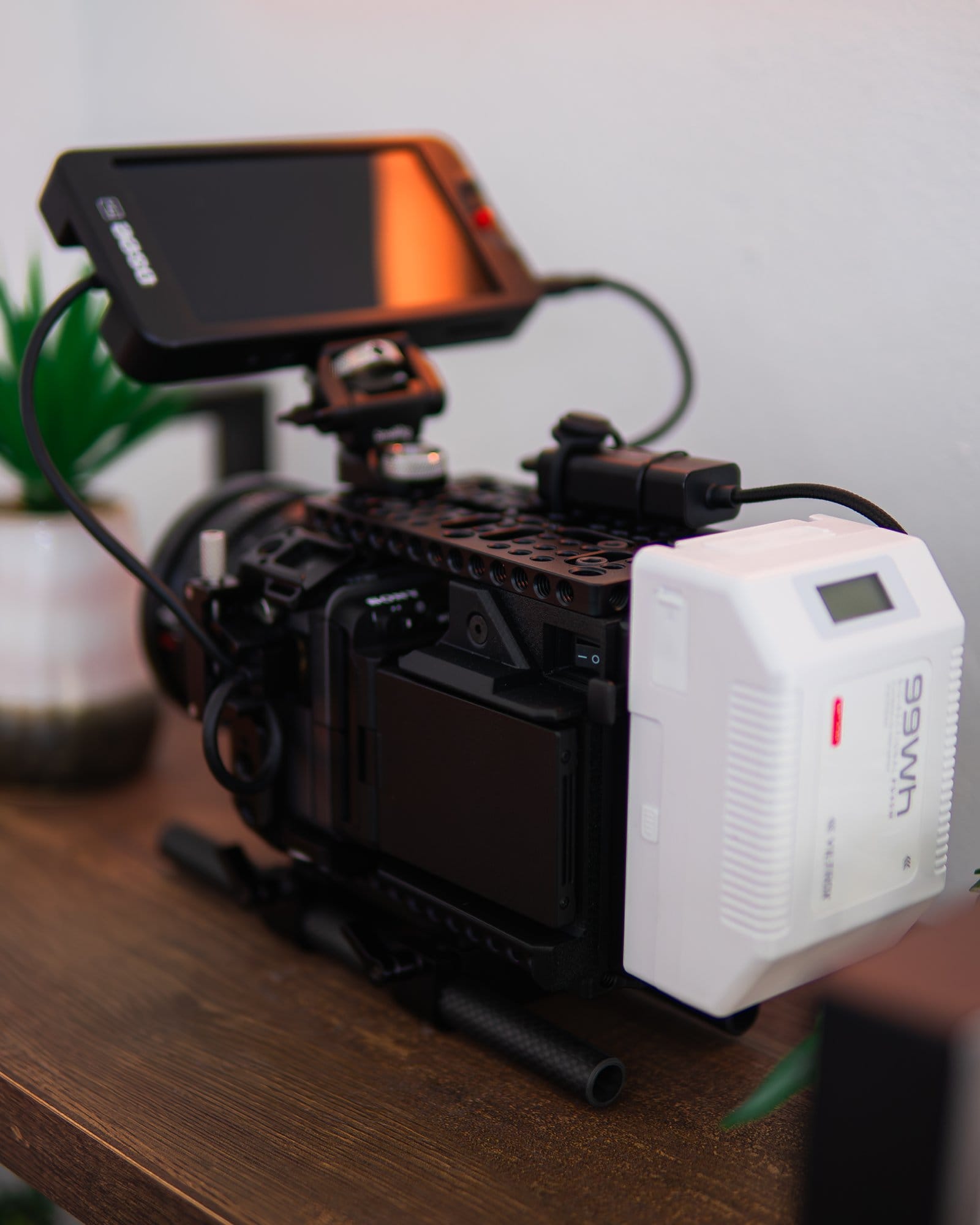 A close-up view of a professional video camera setup, featuring a monitor mounted on top, various cables connected, and a white battery pack attached to the back