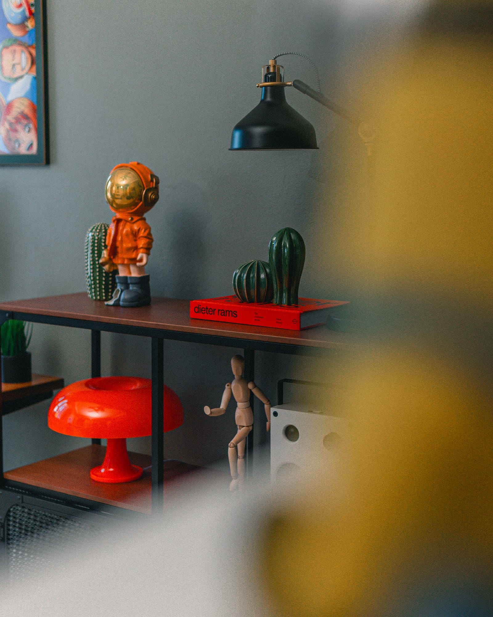 A stylish shelf setup featuring a black lamp, a red Dieter Rams book, green cactus decor pieces, an orange astronaut figurine, an orange mushroom lamp, a wooden mannequin, and a white speaker, with a partial view of a One Piece anime poster in the background